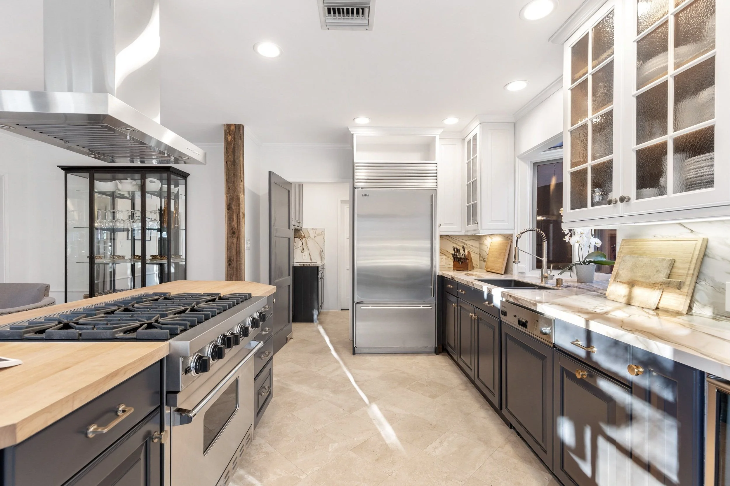 Modern kitchen with black cabinets, marble countertops, stainless steel appliances including a refrigerator and oven, and a wooden kitchen island with a gas stove. There's a glass cabinet with glassware and a potted plant on the counter. Recessed lig