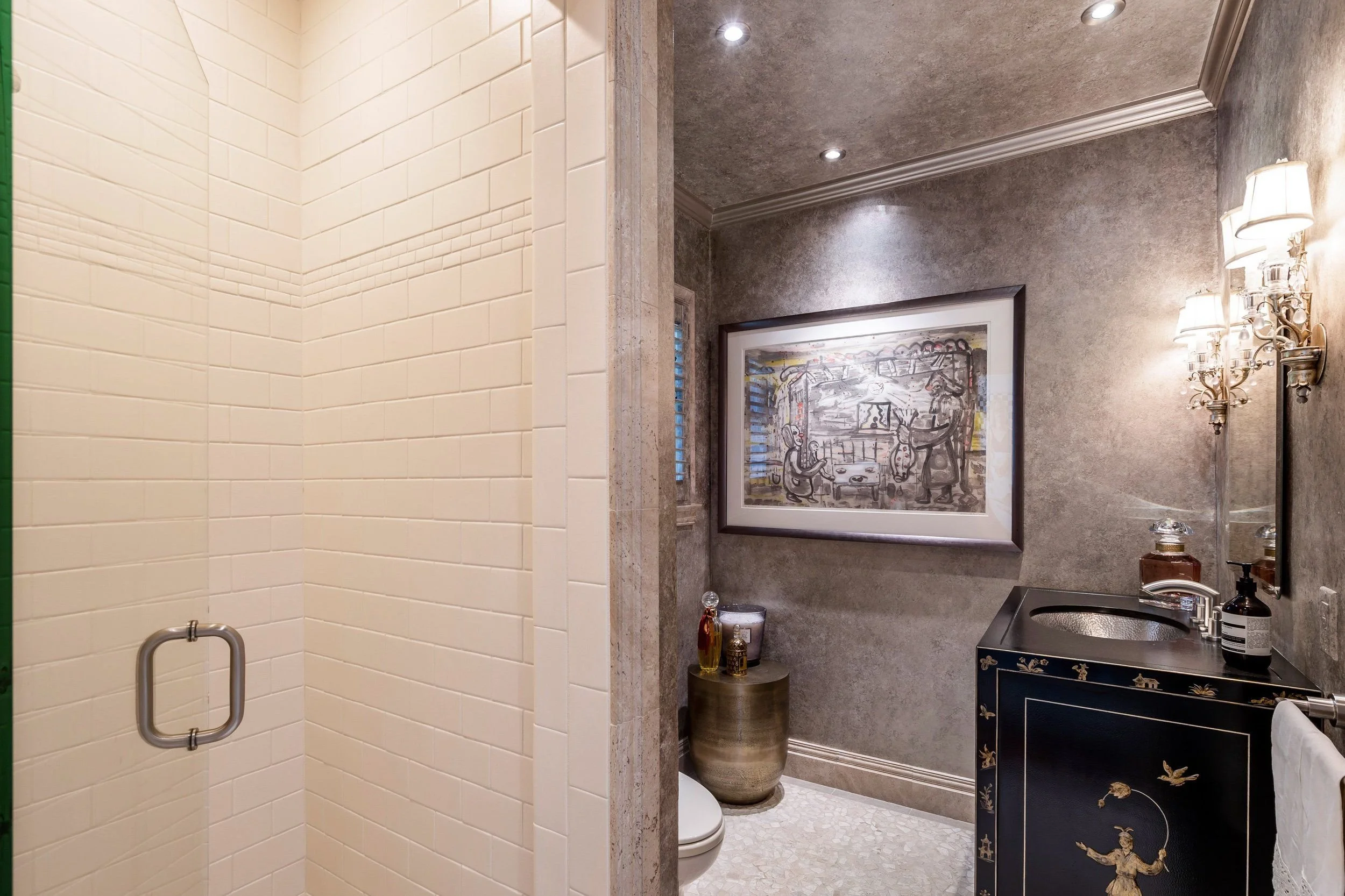 A bathroom with a walk-in shower with cream tiles on the left, a vanity with a black and gold design, a framed artwork on the gray wall, and a wall-mounted sconce with three lamps.