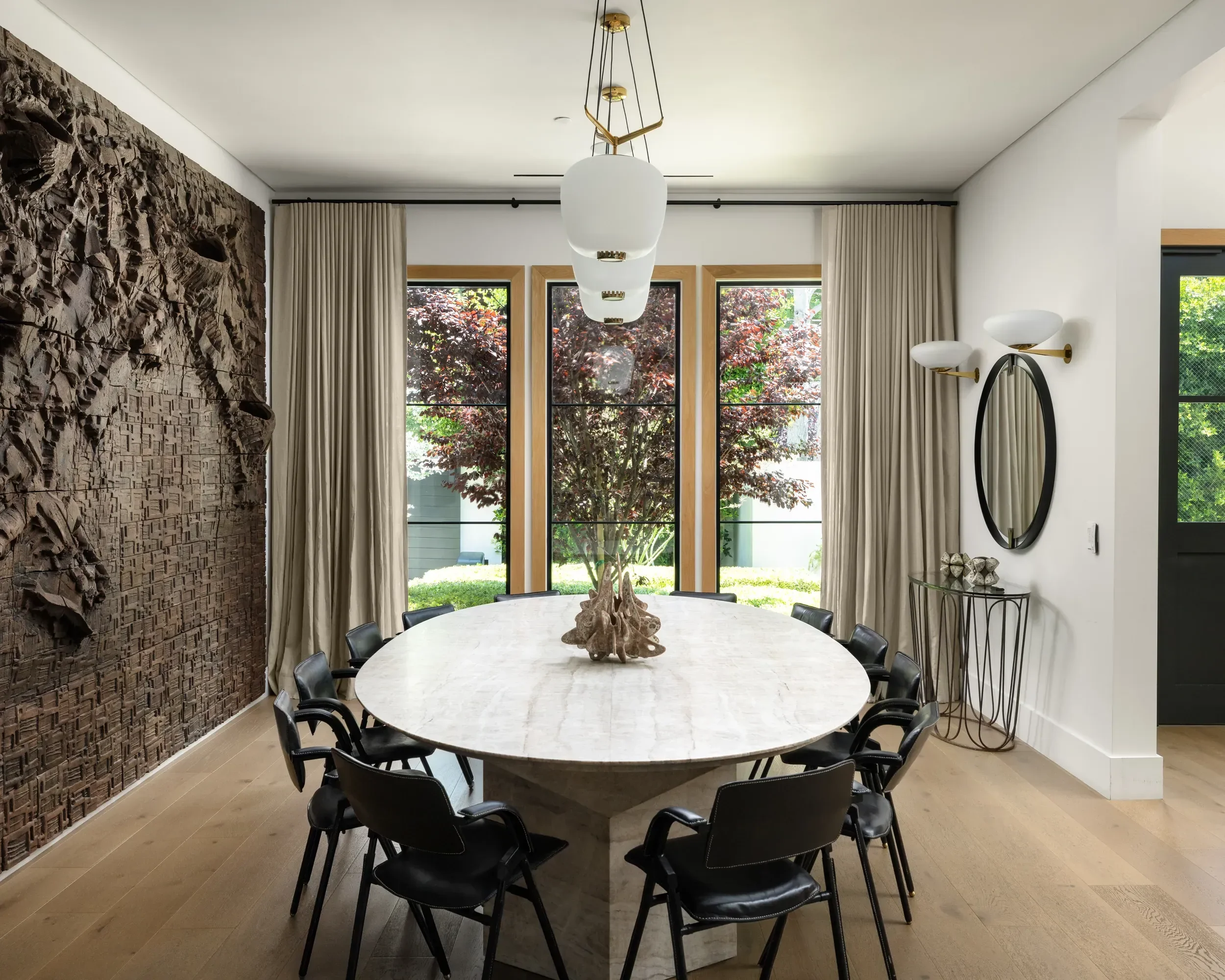 Modern dining room with a large white marble table surrounded by black chairs, a textured wooden wall on the left, large windows with curtains showing trees outside, a contemporary chandelier above the table, a round mirror with wall-mounted light fi