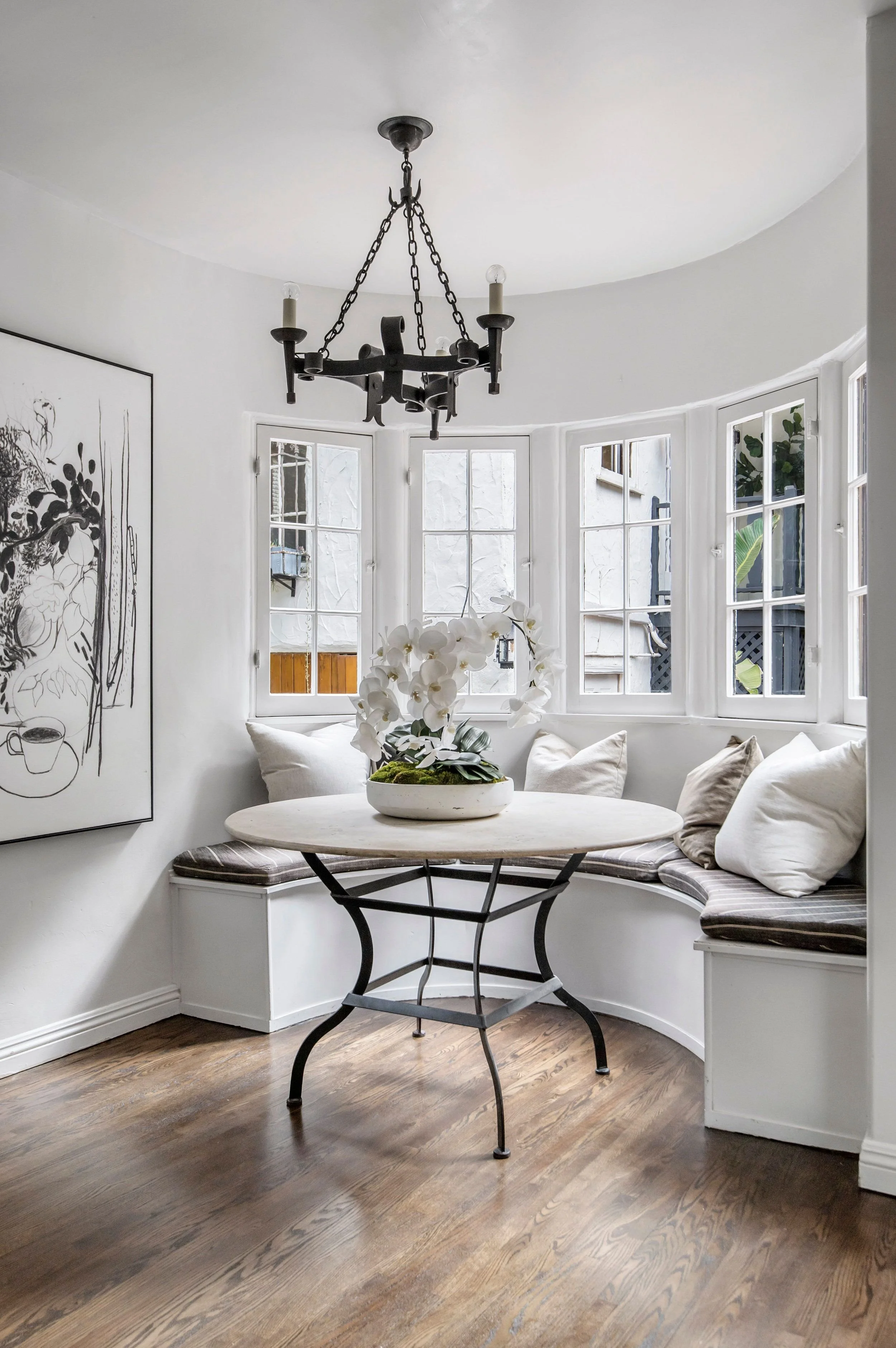 A cozy dining nook with a round table, cushioned bench seats with pillows, a potted white orchid on the table, and a large window with multiple panes and a view of an exterior wall and greenery, all illuminated by natural light, with a black wrought 