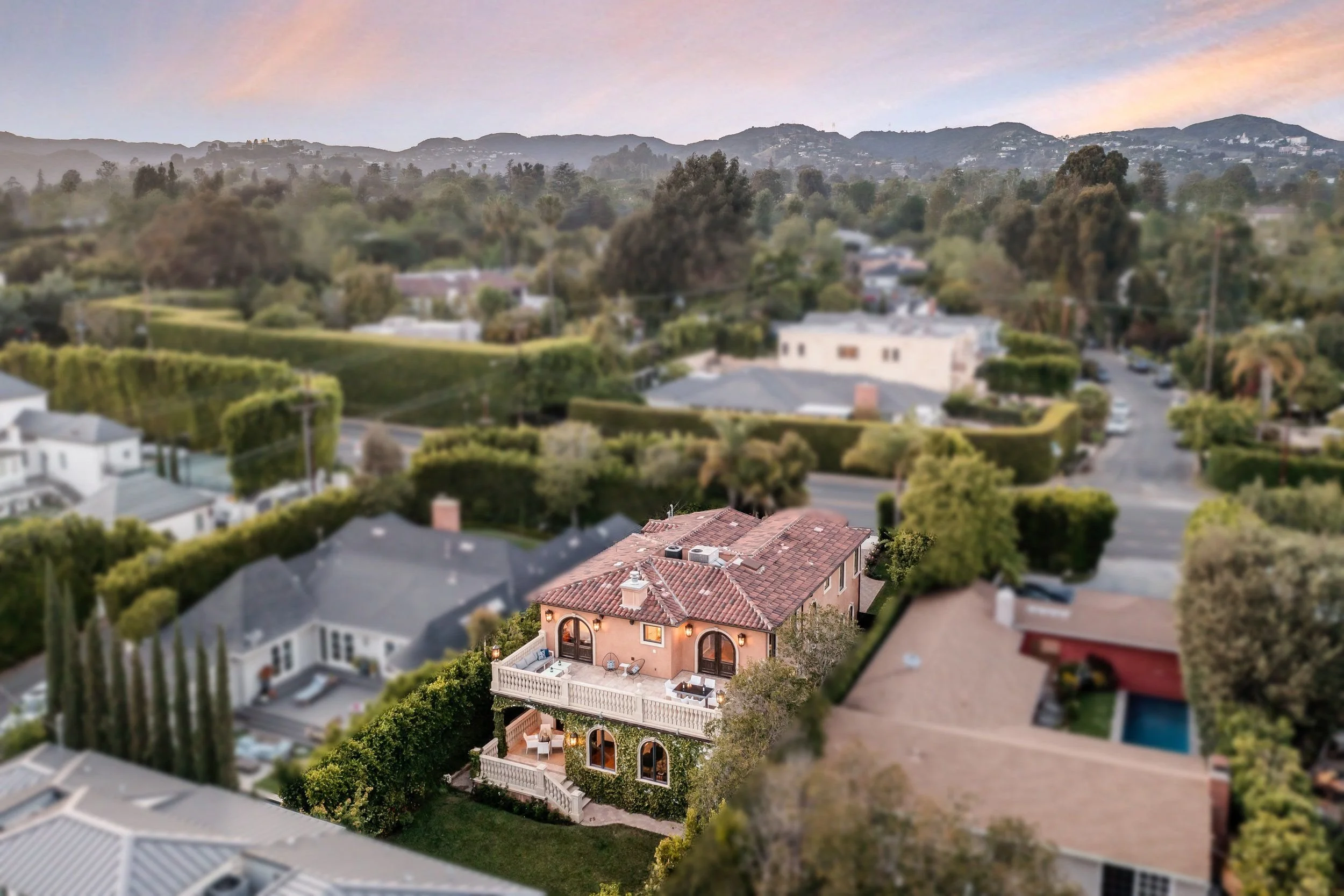 A view of suburban houses with a large, pink Mediterranean-style house in the foreground, surrounded by trees and greenery, with mountains in the distance and a pastel-colored sky.