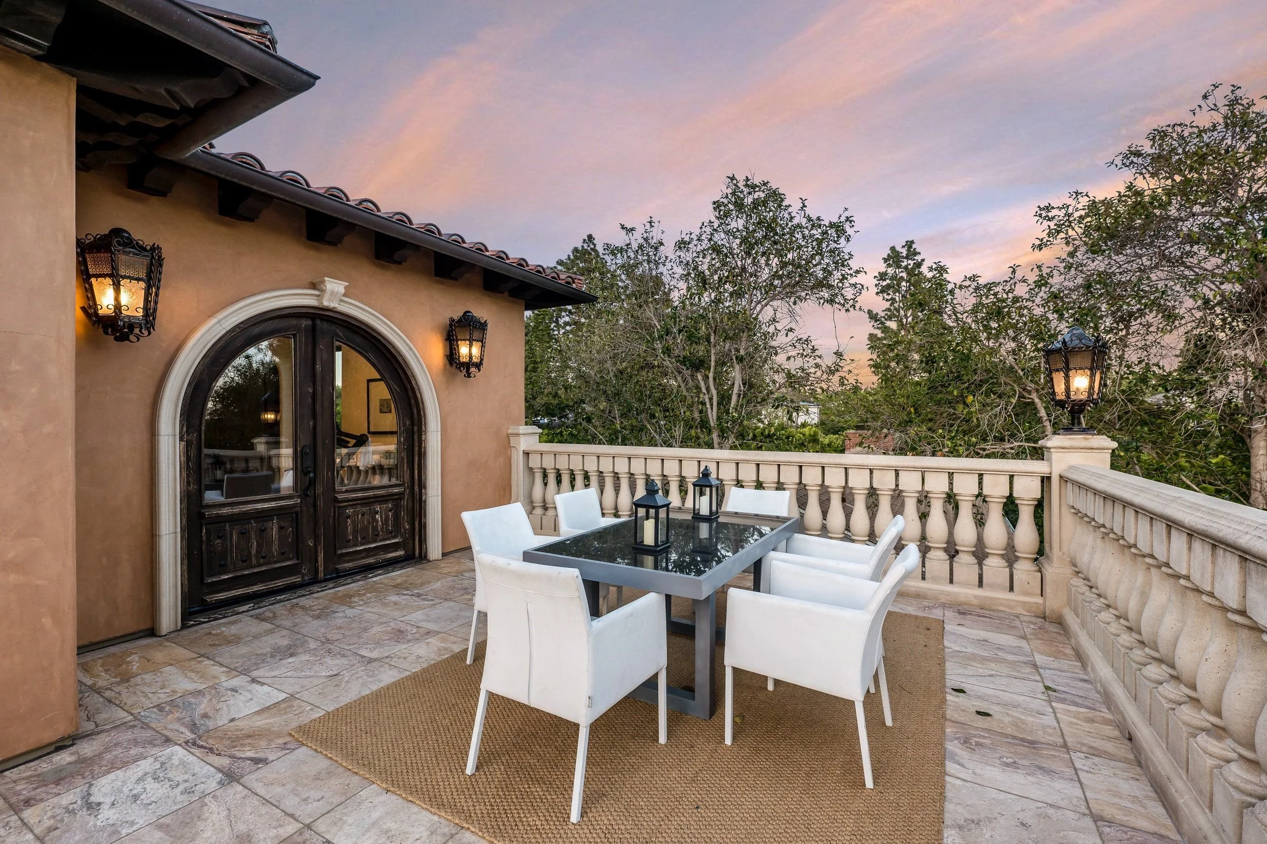 A patio area with a glass table surrounded by white chairs, outdoor wall lamps, and a stone railing, overlooking a lush garden with trees and a colorful sunset sky.
