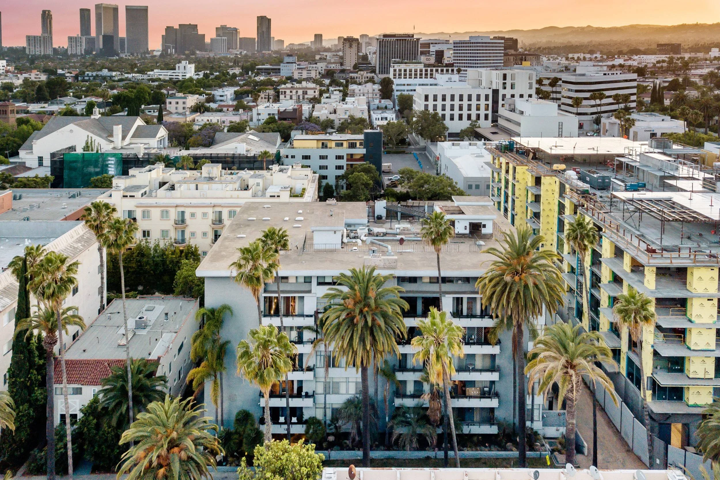 Sunset over a cityscape with tall buildings, residential houses, and palm trees, including a construction site with scaffolding and building materials in the foreground.