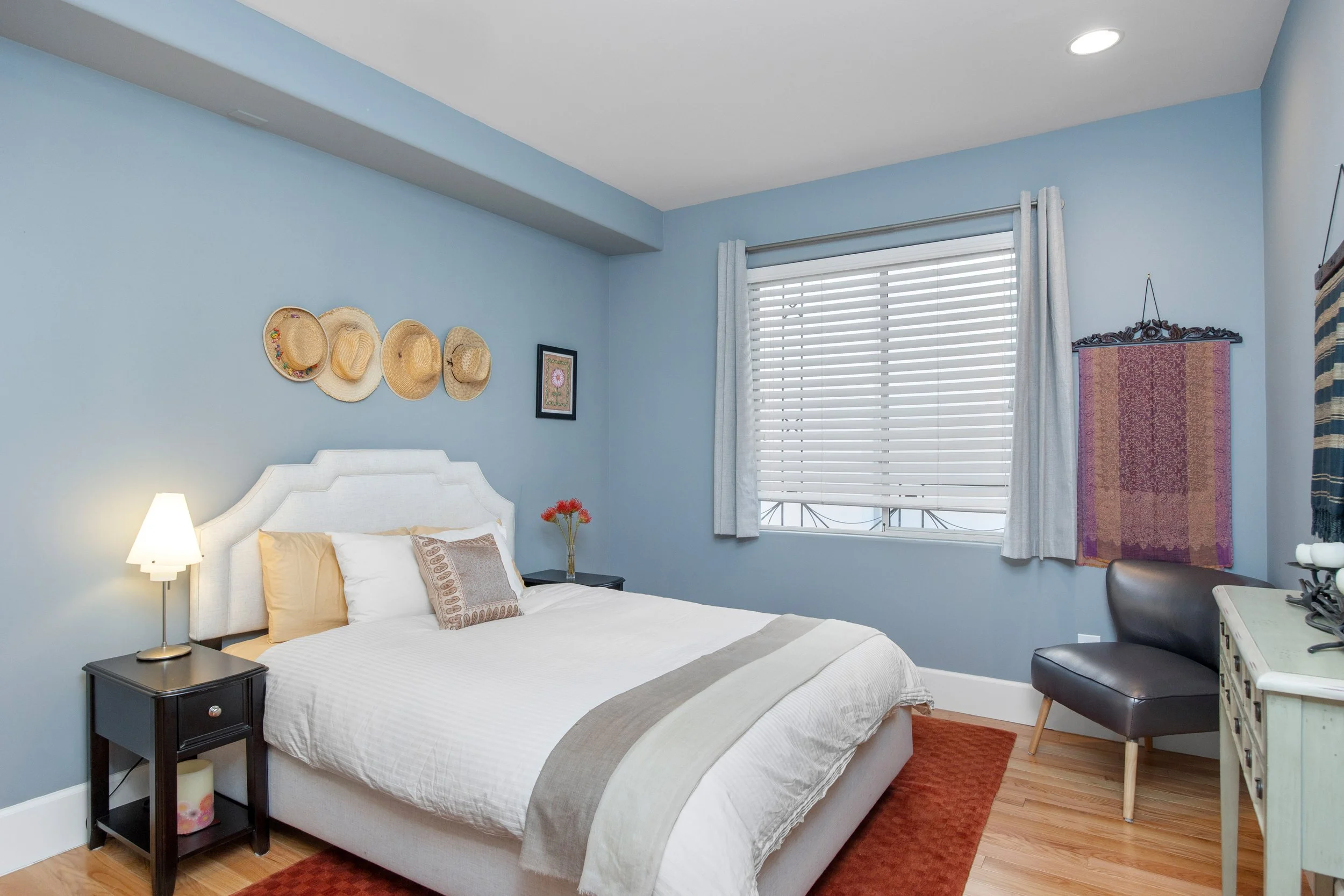 A bedroom with light blue walls, a white bed with beige and patterned pillows, a black nightstand with a lamp, a window with white blinds and curtains, decorative straw hats on the wall, framed art, and a hanging tapestry.