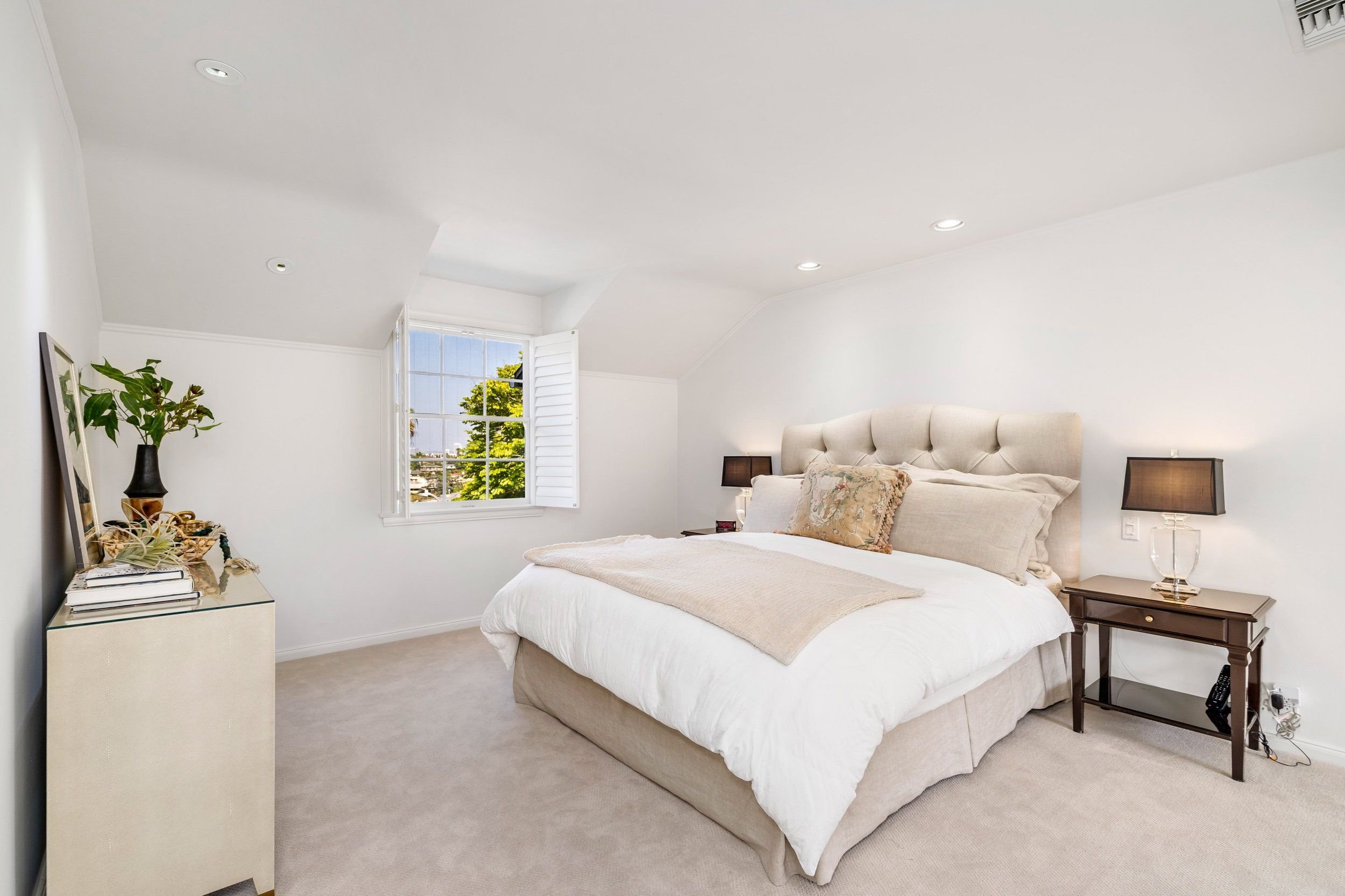 A bedroom with a large beige bed, beige bedding, and pillows. A window with white shutters shows a tree outside. There are nightstands with lamps on each side of the bed. A dresser or side table on the left holds plants, books, and decorative items. 