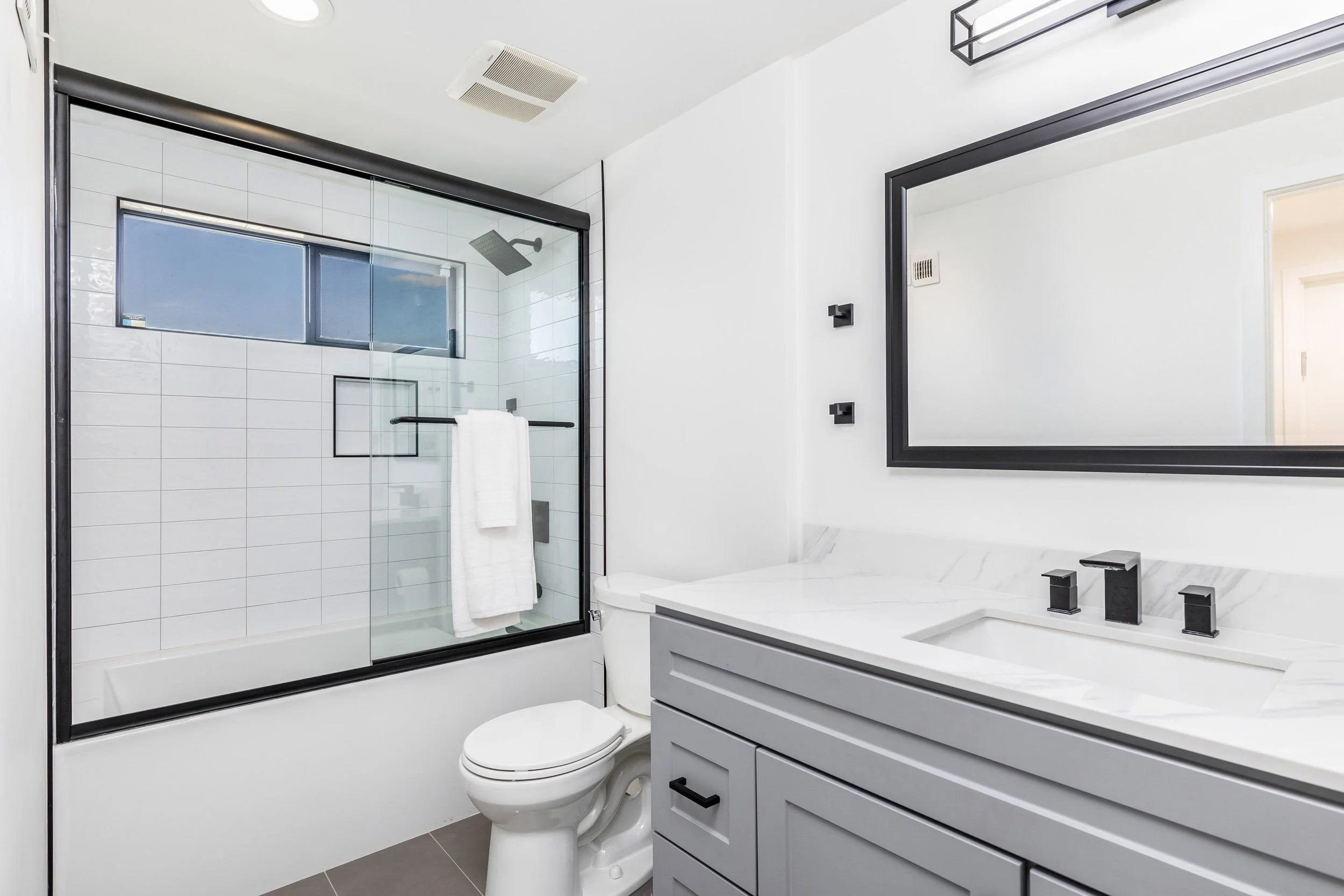 Modern bathroom with a glass-enclosed shower, white tiles, a white toilet, gray cabinetry, and a large mirror.
