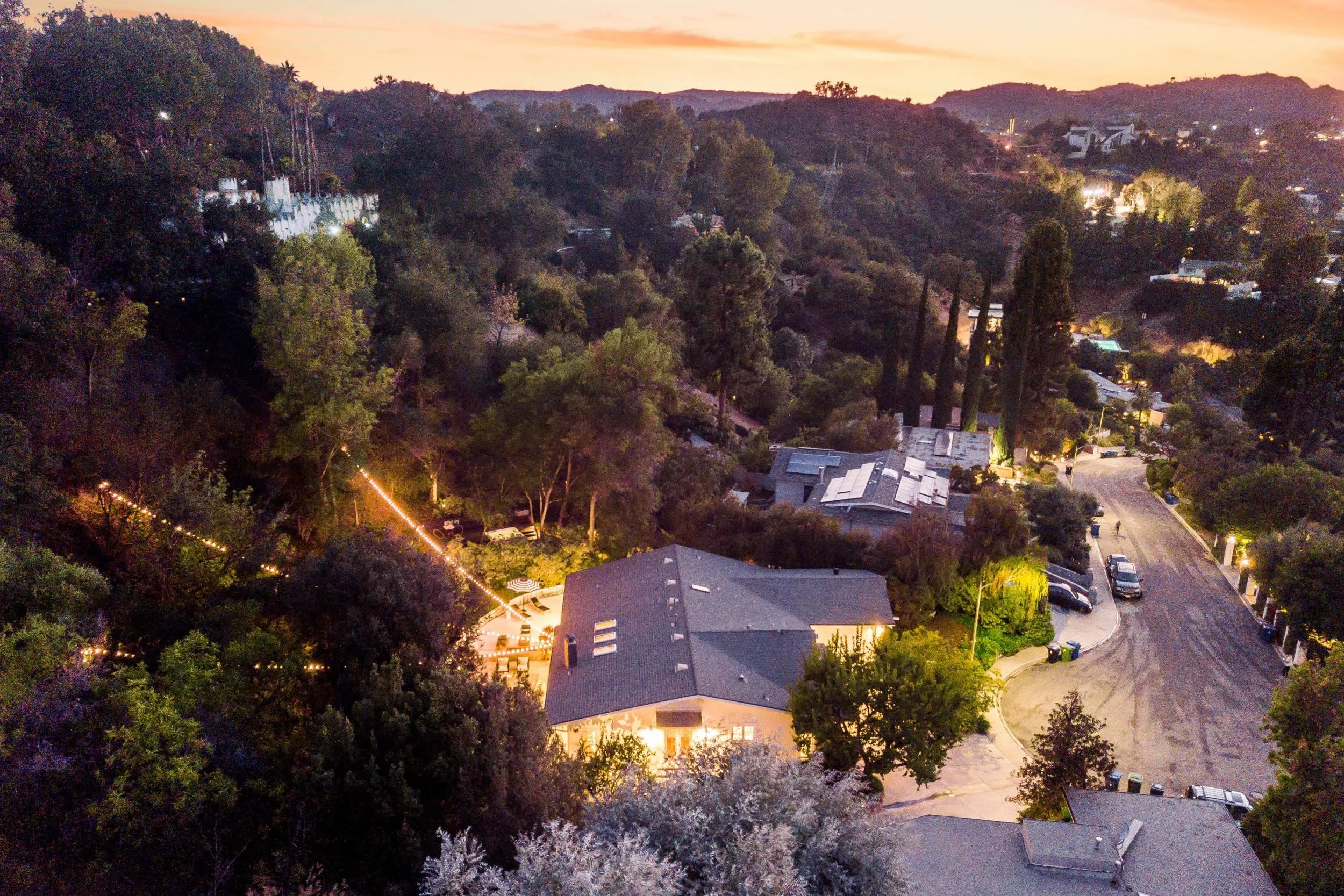 A hillside residential neighborhood at dusk with houses, trees, and outdoor lighting illuminating the area.