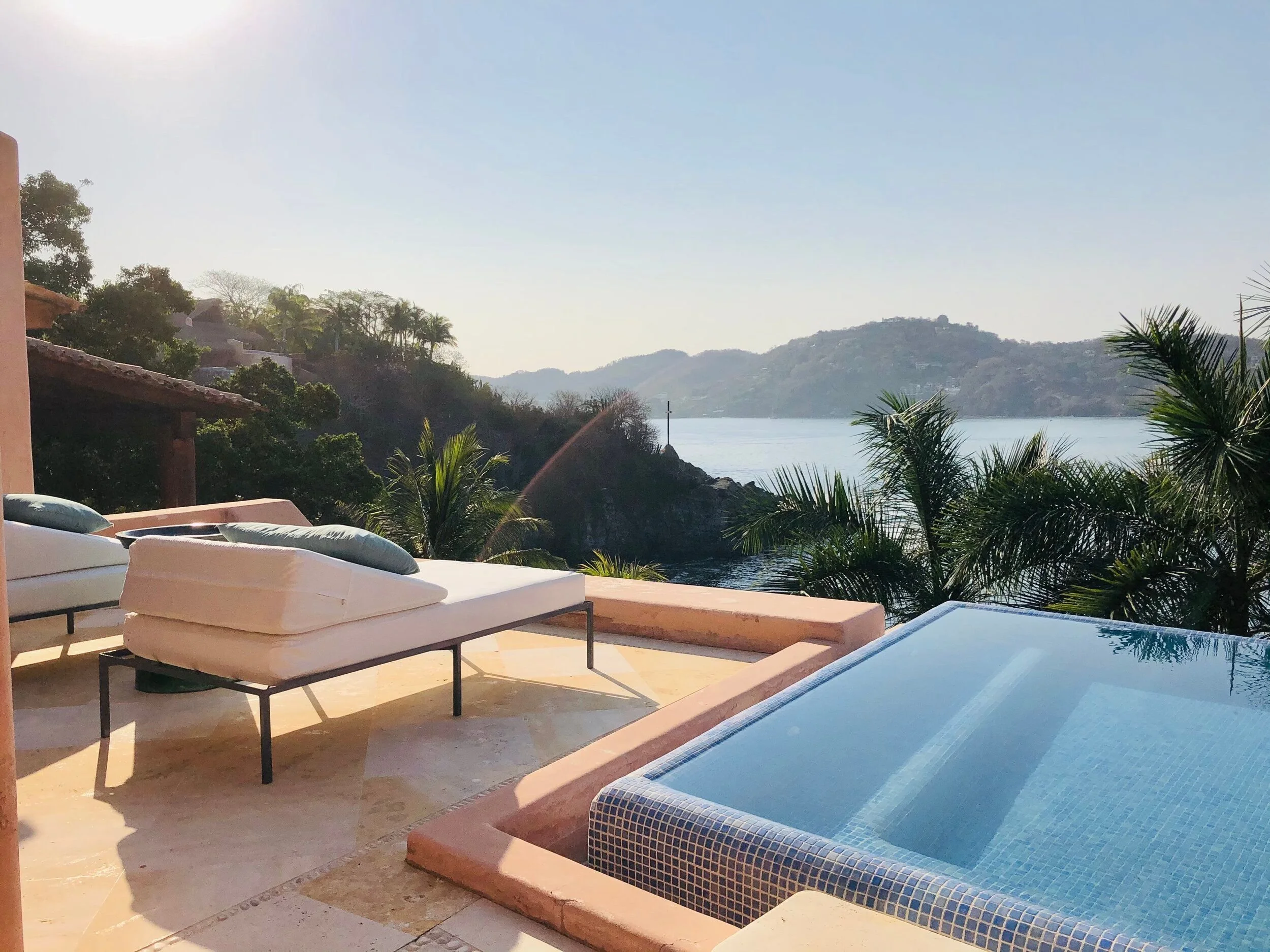 Outdoor lounge area with sunbeds and an infinity pool overlooking a lake or river, surrounded by green hills and trees under a clear blue sky.