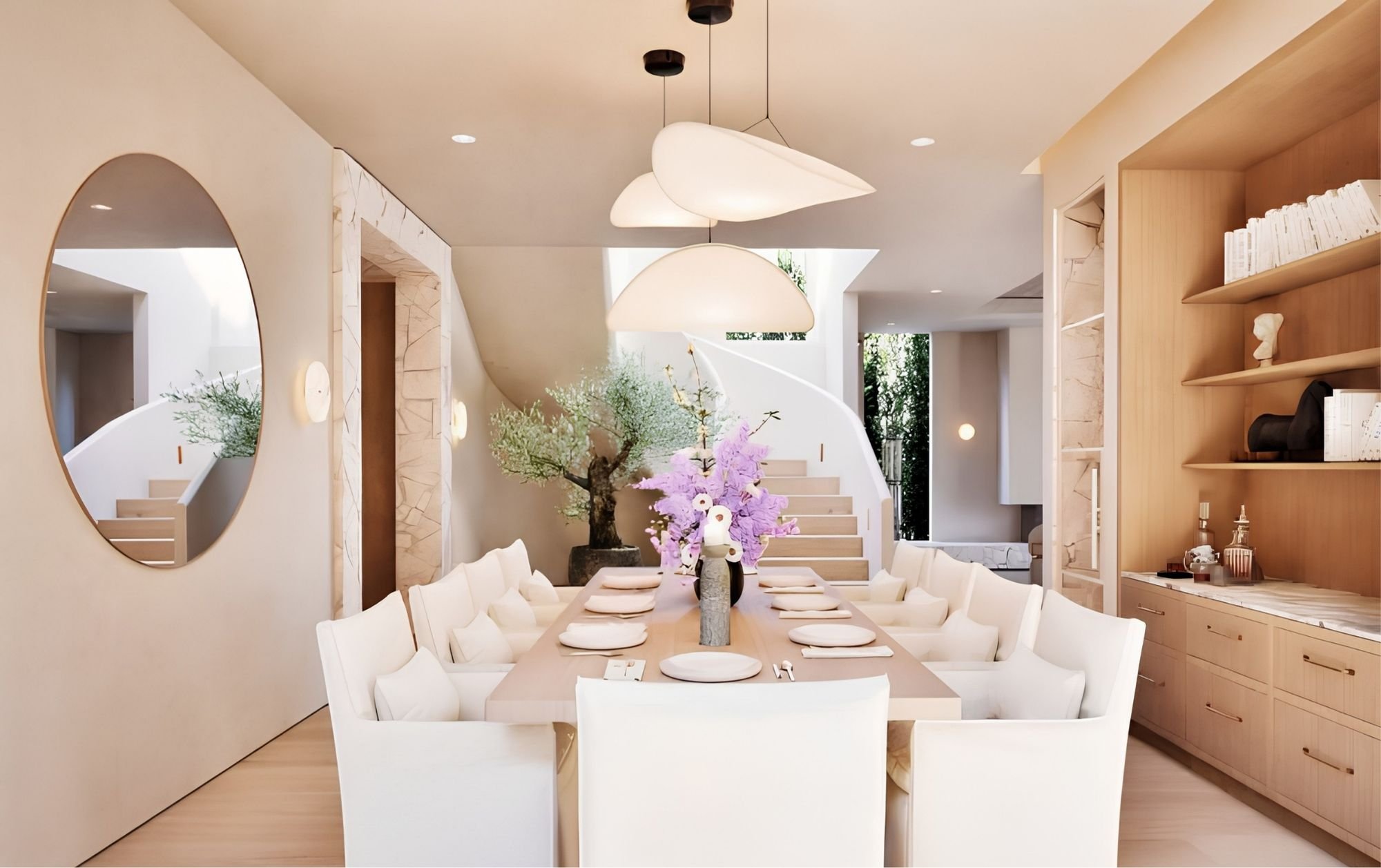 A modern dining room with a long table set with white plates and napkins, surrounded by white chairs, and decorated with a large floral centerpiece. The room features contemporary lighting fixtures, a large round mirror on the wall, and an open space