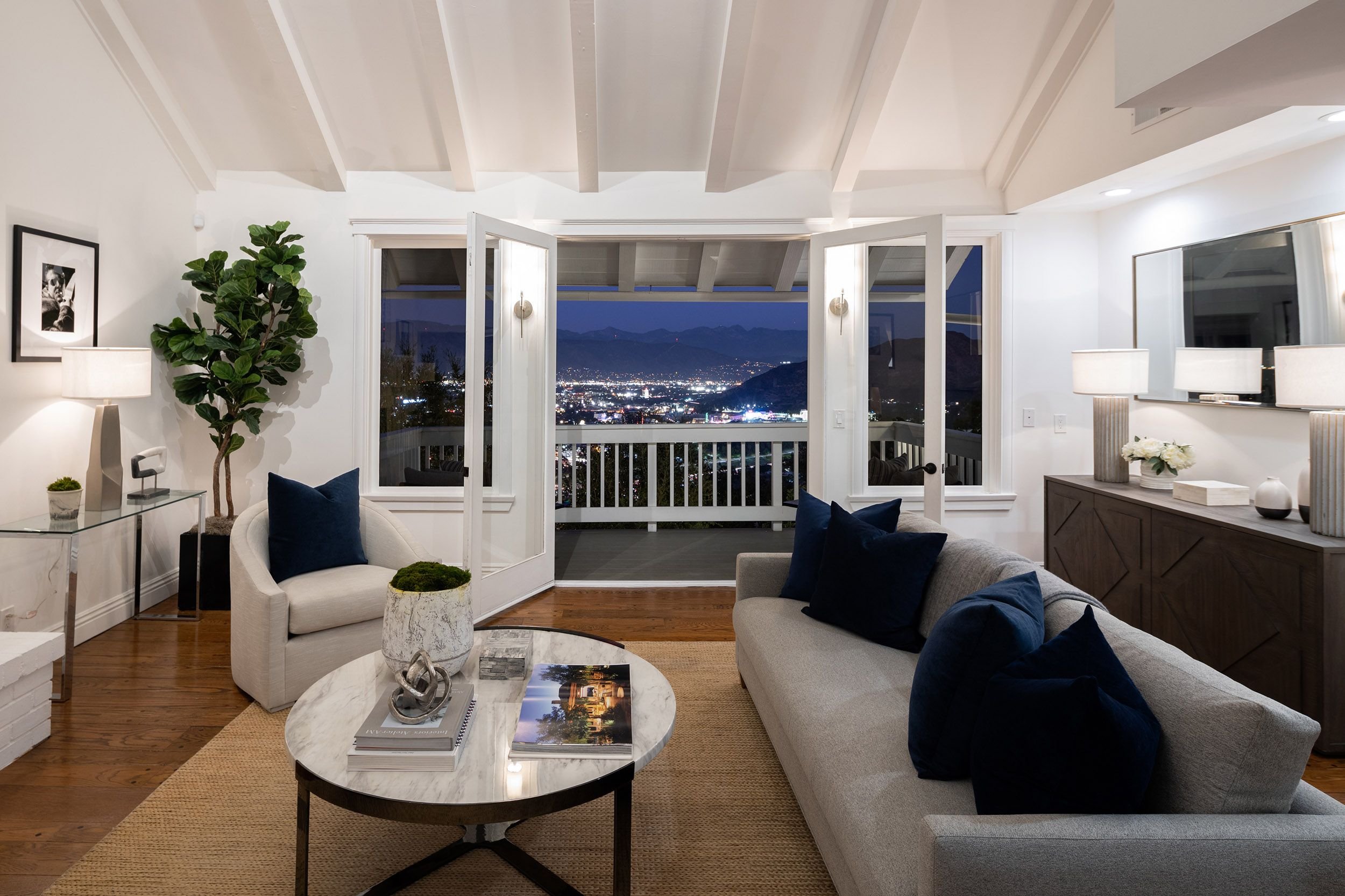 A living room with open balcony doors revealing a nighttime cityscape and mountain view, furnished with white and gray seating, wooden floor, and modern decor.