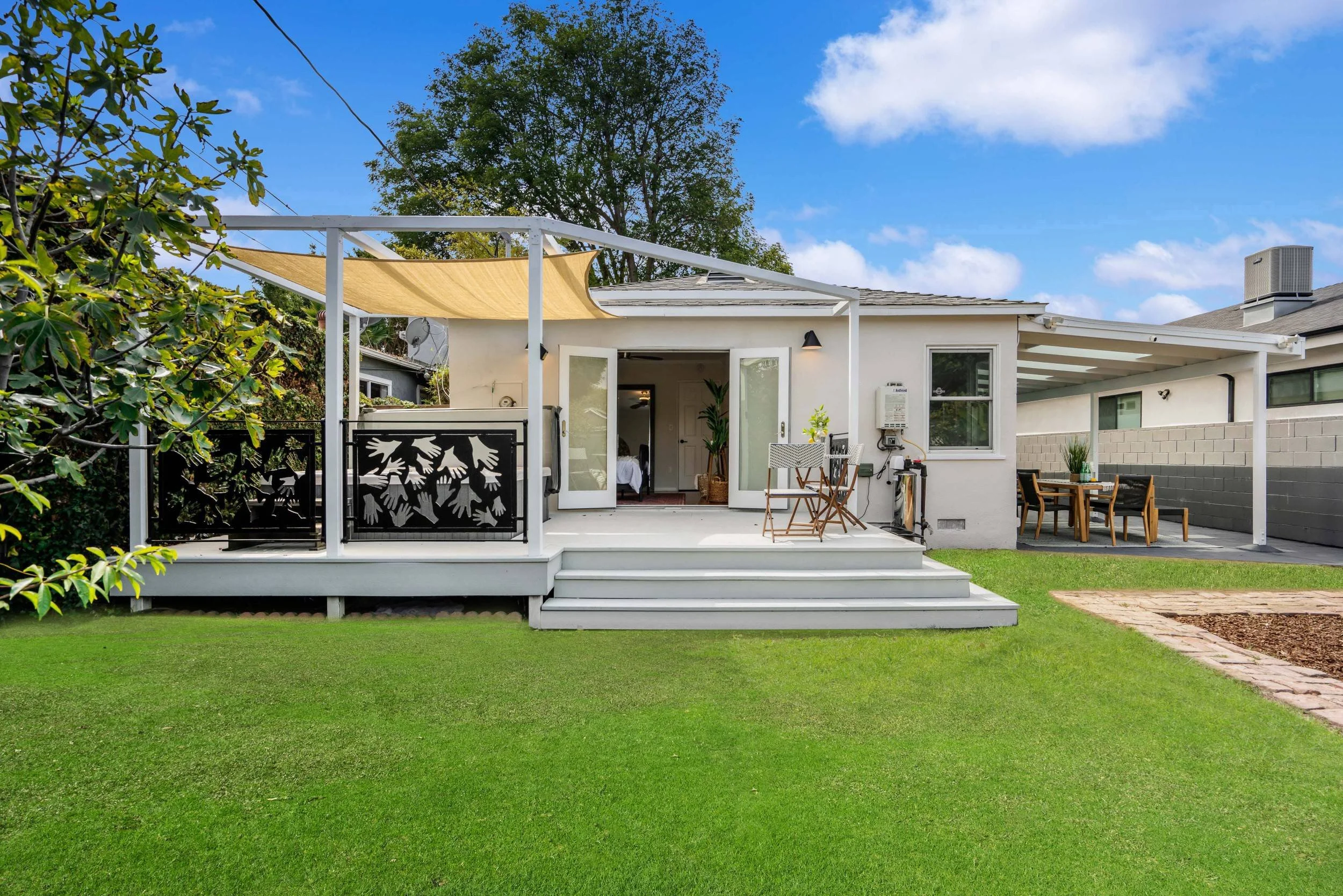 A backyard with a grassy lawn, a white deck with steps, and a white house with a sliding glass door open to the deck. There is outdoor furniture on the deck, including chairs and a table. A beige shade sail provides cover over part of the deck. The s