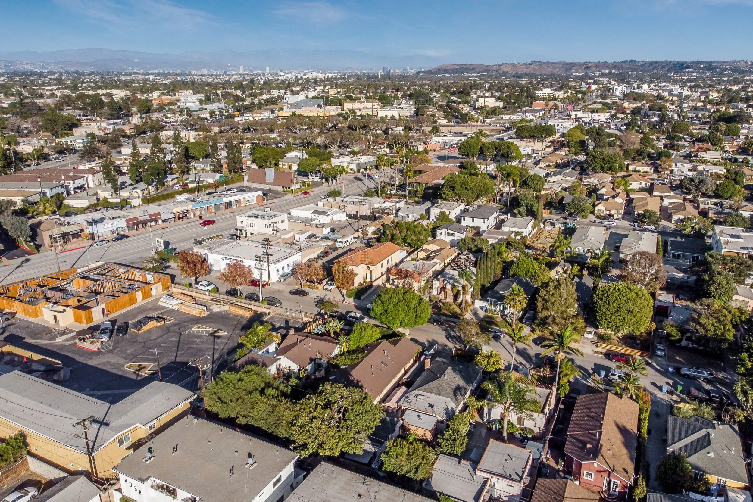 Aerial view of a cityscape showing numerous residential houses, trees, a commercial strip mall with parking lot, and distant mountains under a blue sky.