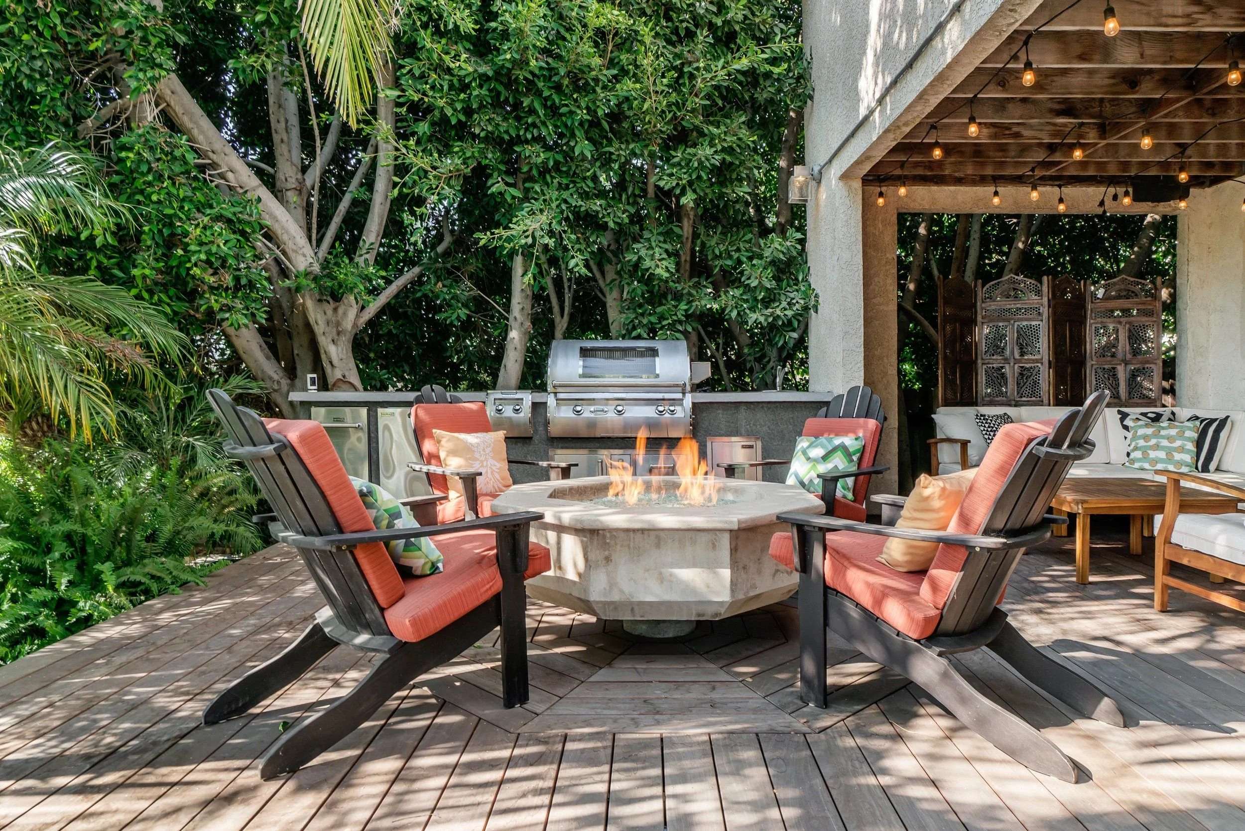 Outdoor patio with a fire pit at the center surrounded by chairs with cushions, a grill, a seating area with pillows, string lights hanging from the ceiling, and lush green trees and plants surrounding the space.