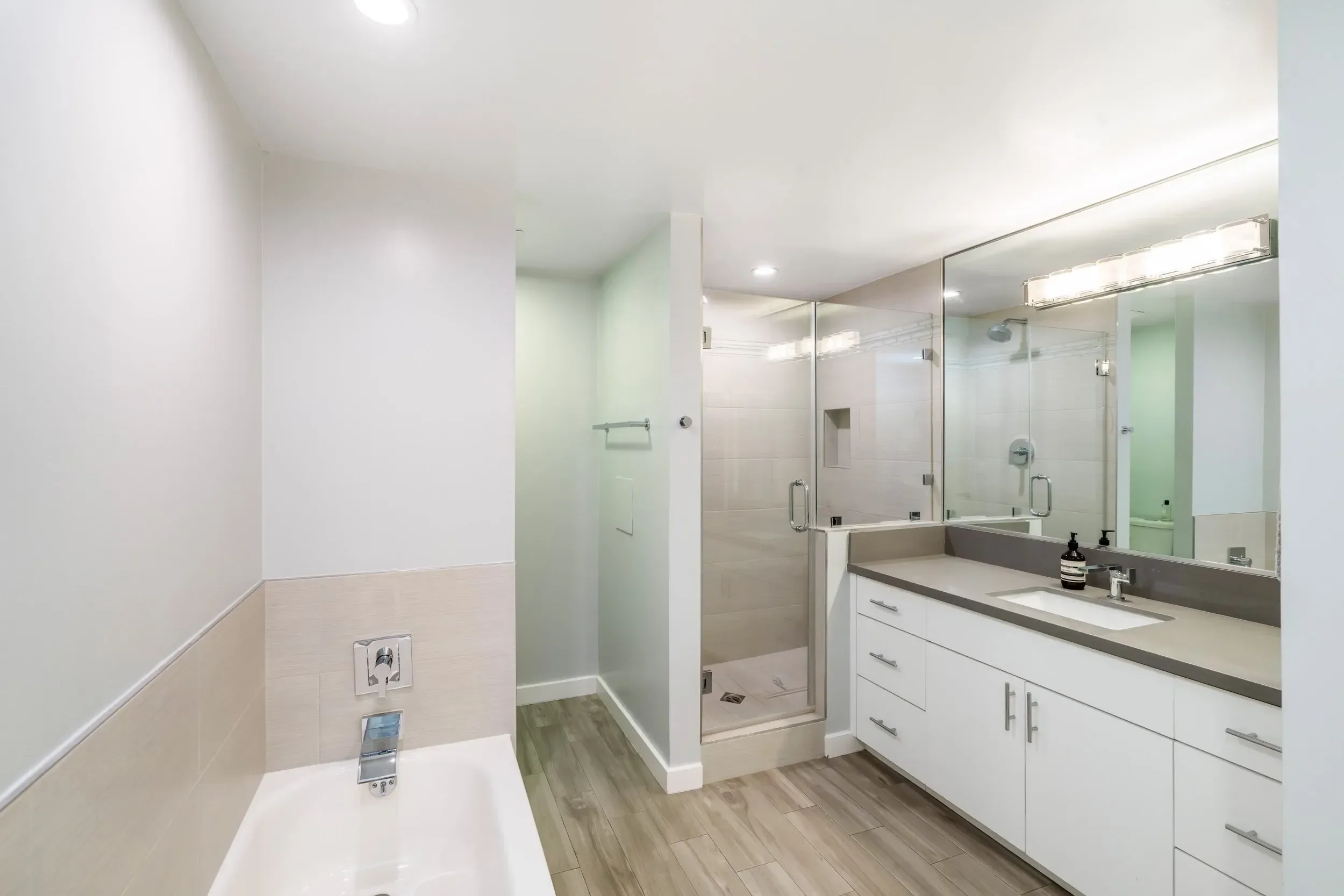 A modern bathroom featuring a shower with glass doors, a large mirror above a white vanity with drawers, a granite countertop, and a sink with a black-striped soap dispenser. The room has white and beige tile walls, wood-like flooring, and bright cei