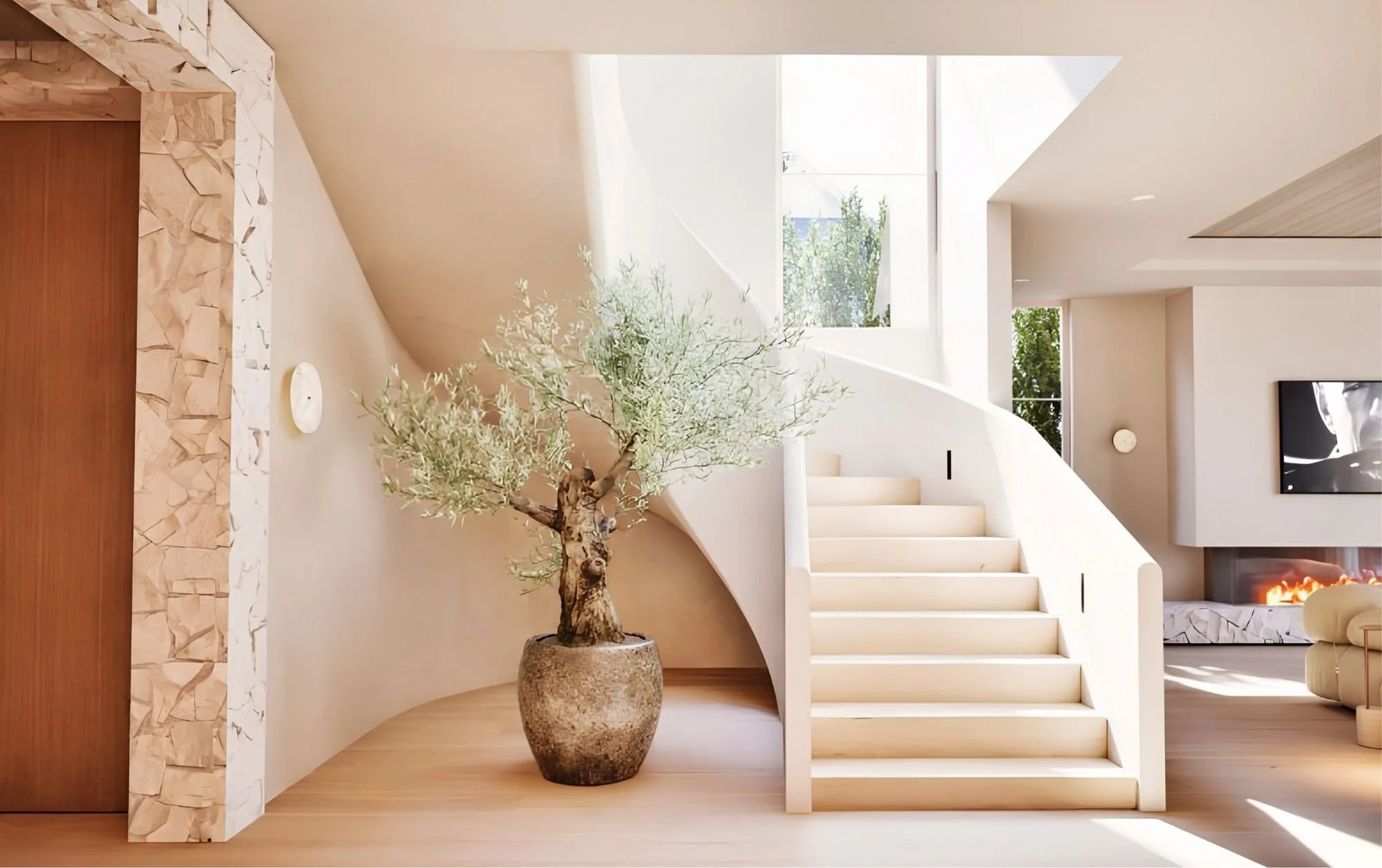 Interior of a modern house with wooden stairs, a large potted olive tree, and a fireplace with a fire burning.