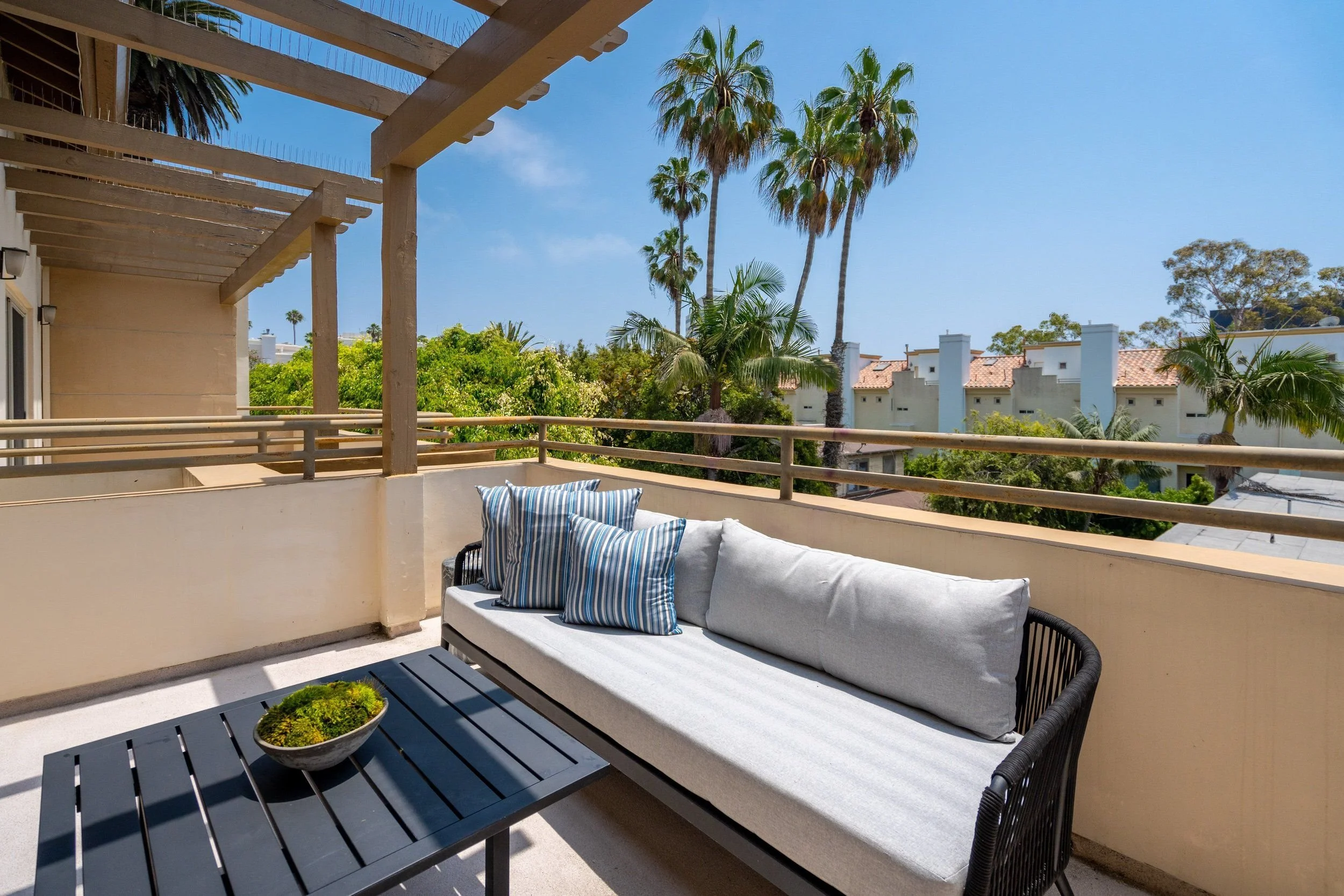 Balcony with black sofa and blue-striped pillows, black coffee table with a bowl of green plants, beige railings, white walls, and a wooden trellis. Background shows palm trees and a clear blue sky.