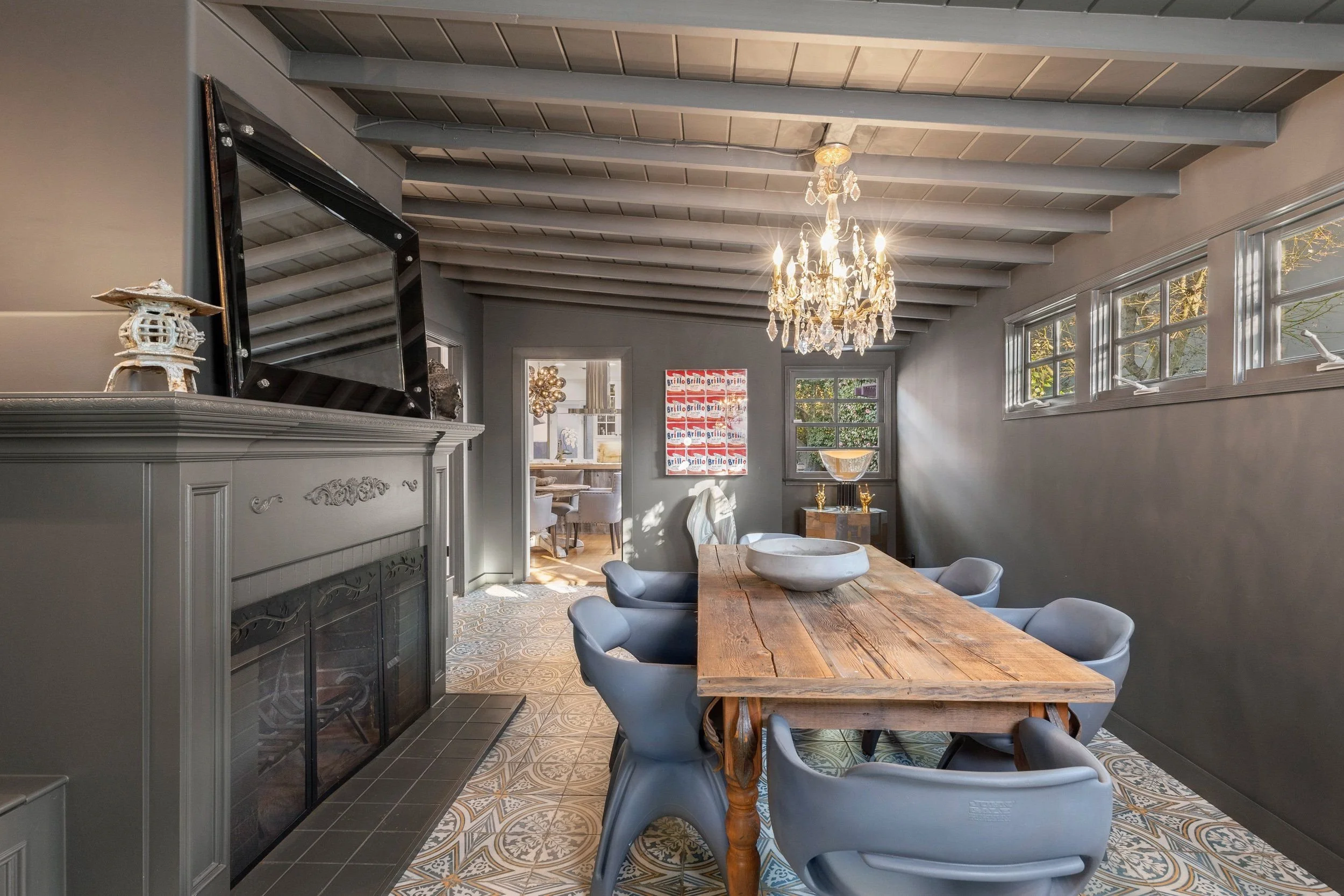 A dining room with a wooden table surrounded by six gray chairs, a crystal chandelier hanging from the ceiling, and a windowed side wall with decorative art on the wall.