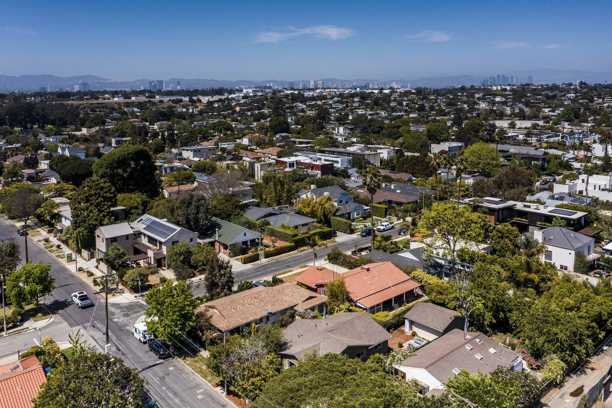 Aerial view of a suburban neighborhood with houses, trees, and streets, with city skyline and mountains in the distance under a clear blue sky.