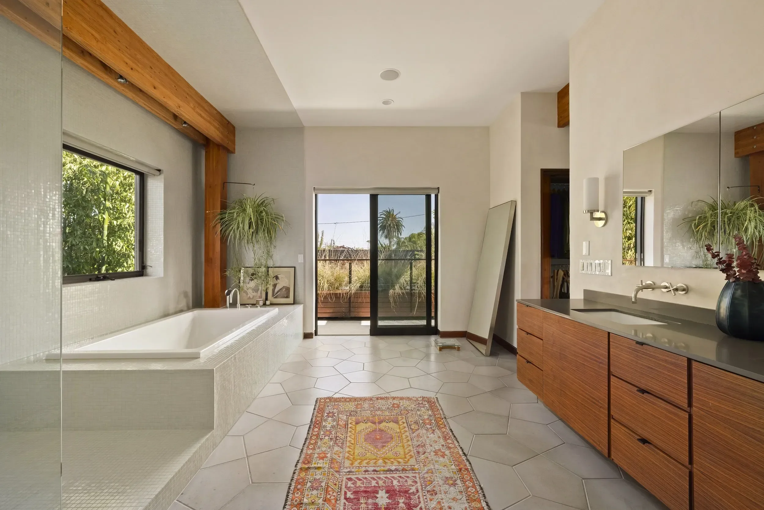 A modern bathroom with a built-in bathtub, large windows showing outside greenery, wooden accents, a bathroom vanity with a sink, a mirror, a small colorful rug, and sliding glass doors leading to an outdoor patio.