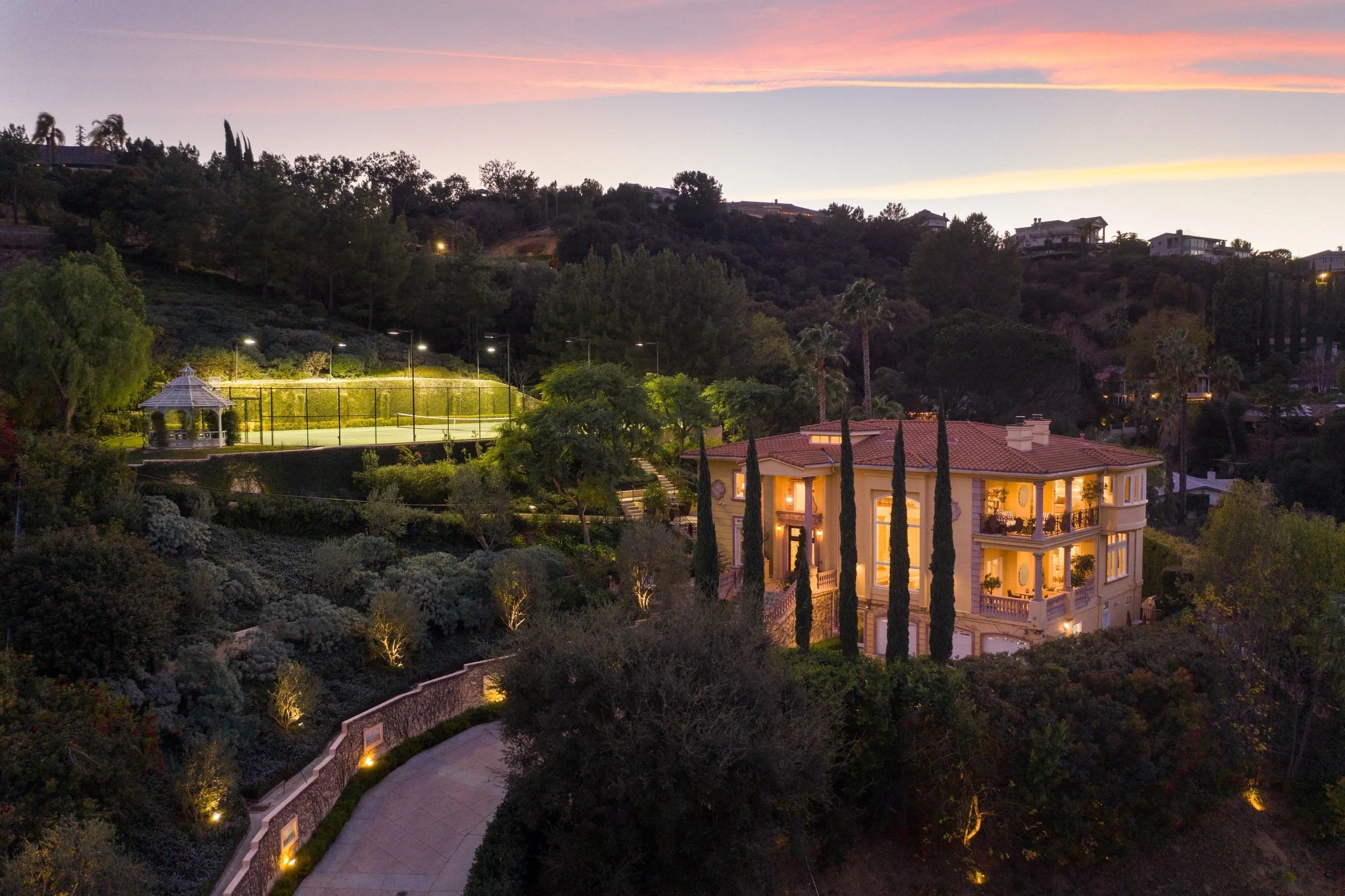 A hillside residential area during dusk featuring a large, elegant house with glowing lights, surrounded by trees and a landscaped yard. In the background, more houses are visible on the hillside, with a tennis court lit up in the foreground.