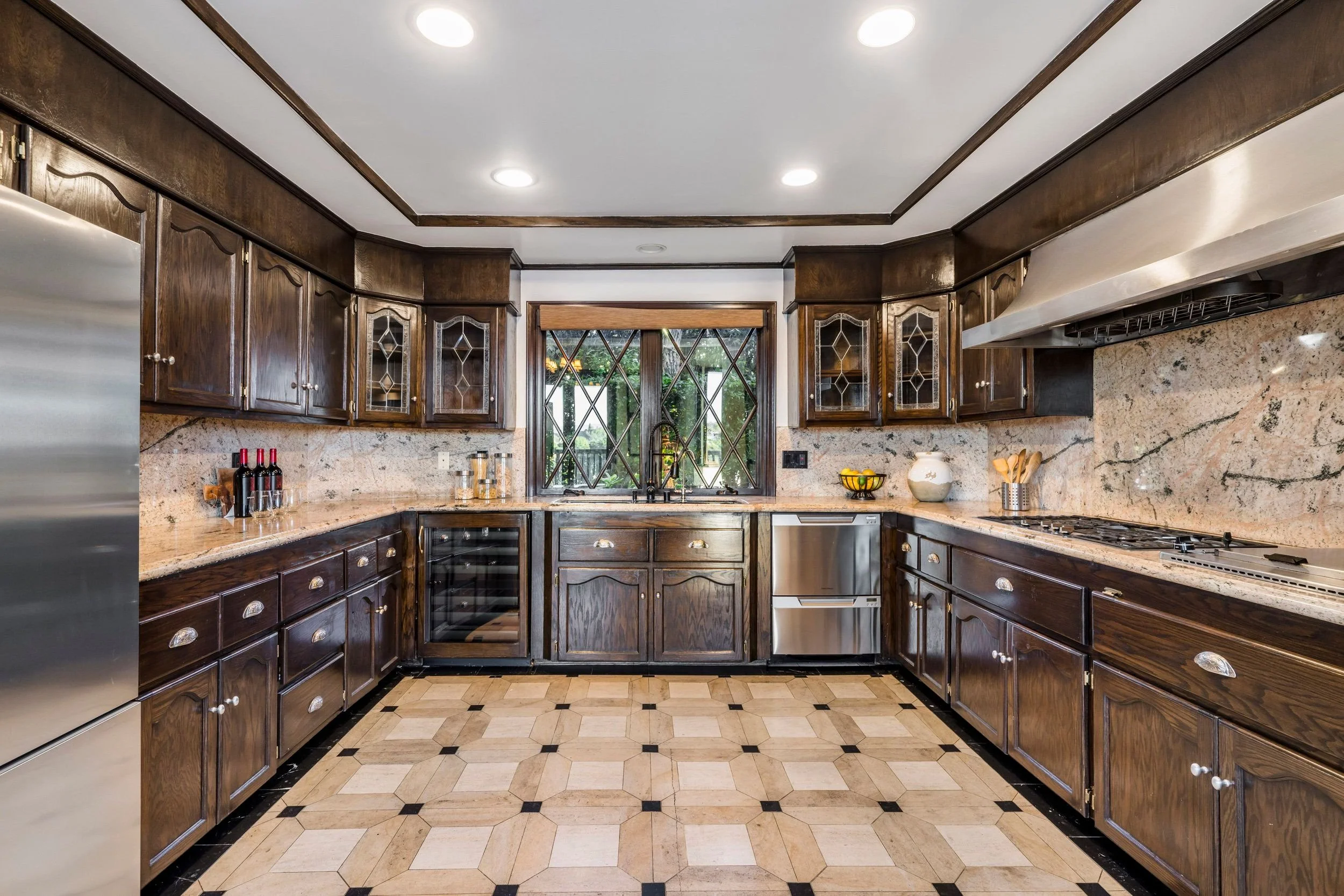 A kitchen with dark wooden cabinets, beige granite countertops, and matching backsplash. There is a window over the sink, a wine cooler, and a stove with a range hood. The floor has a pattern of hexagonal tiles.