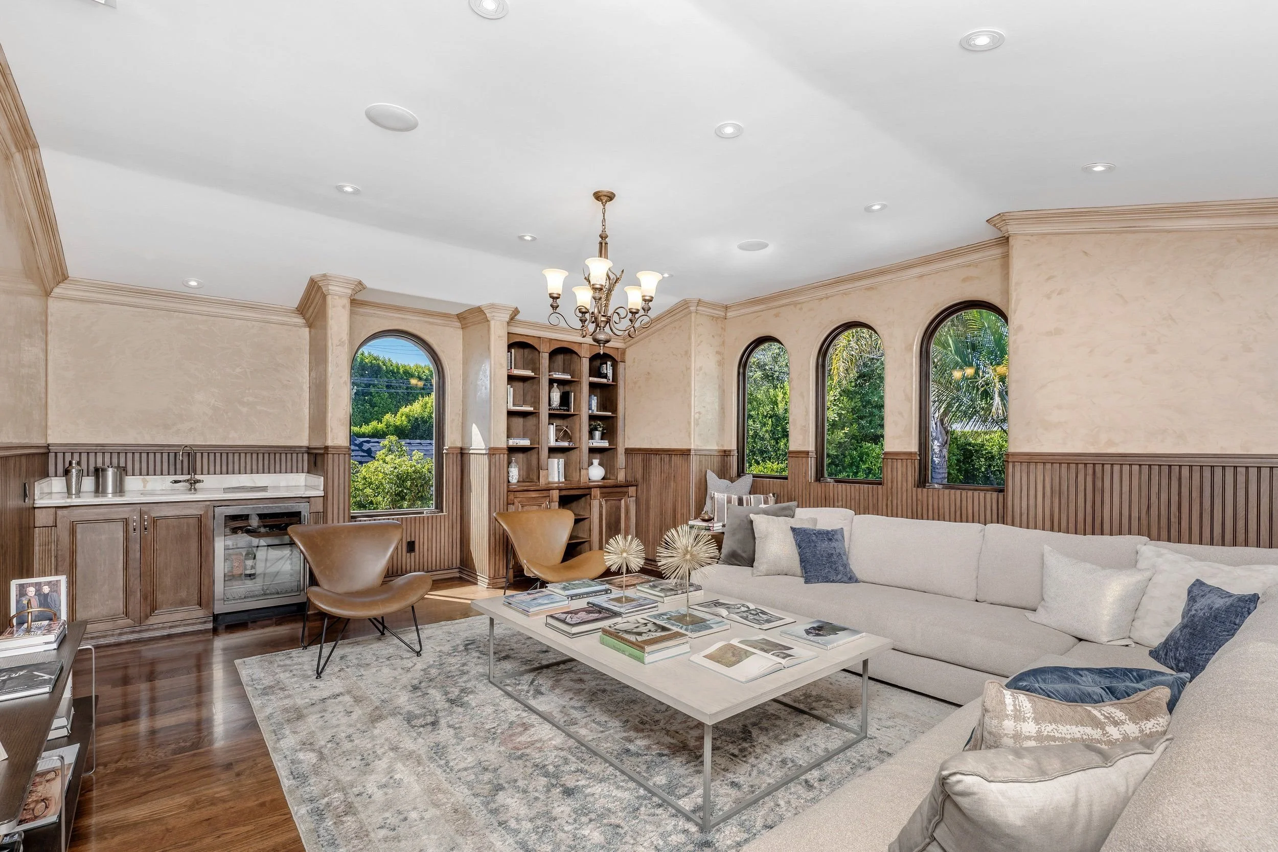 Living room with a white sectional sofa, wooden coffee table, chairs, built-in wooden shelves, large arched windows showing greenery, chandelier, and hardwood floors.