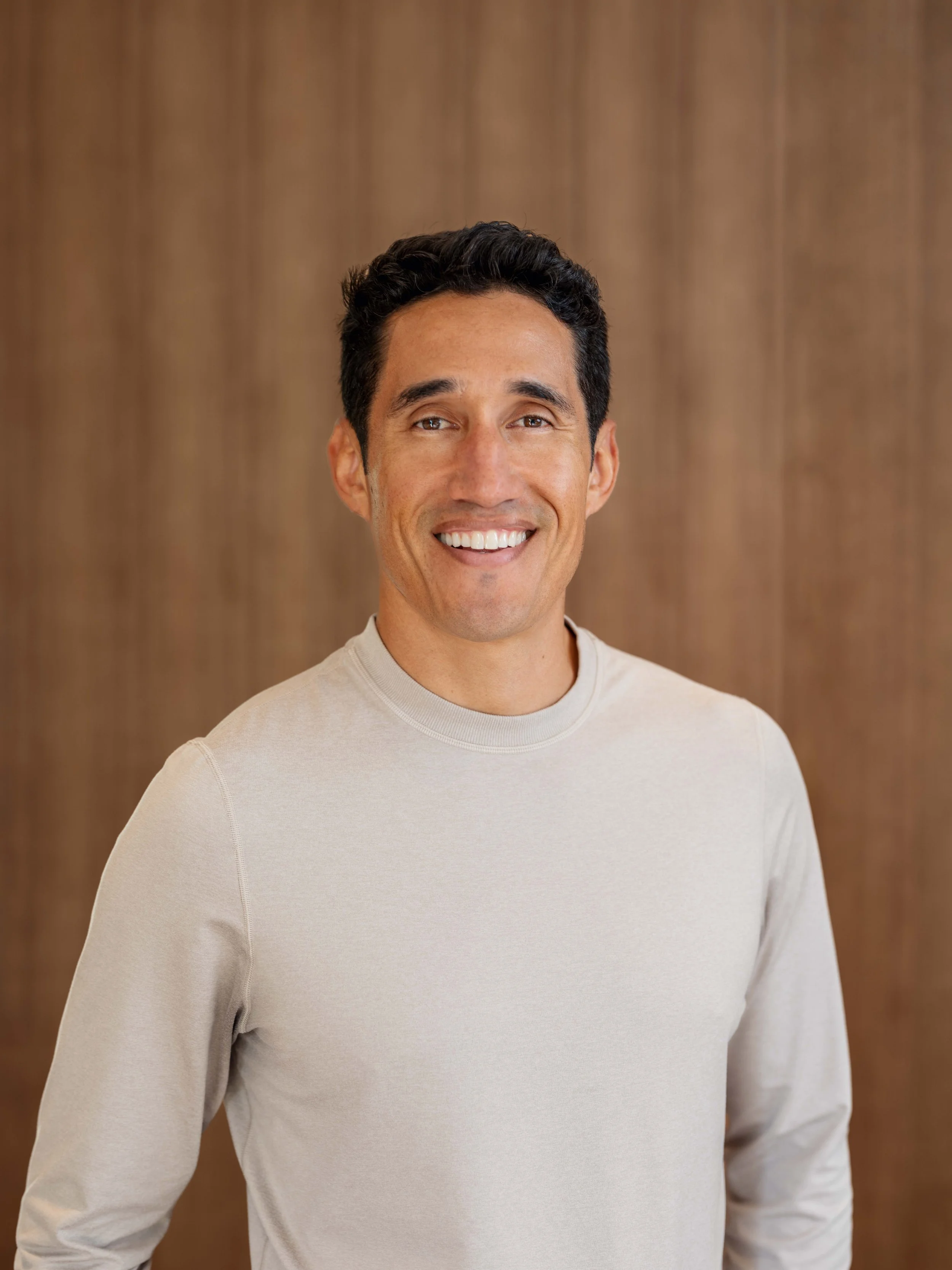 Headshot of a smiling man with dark hair, wearing a beige long-sleeve shirt, standing in front of a wooden background.