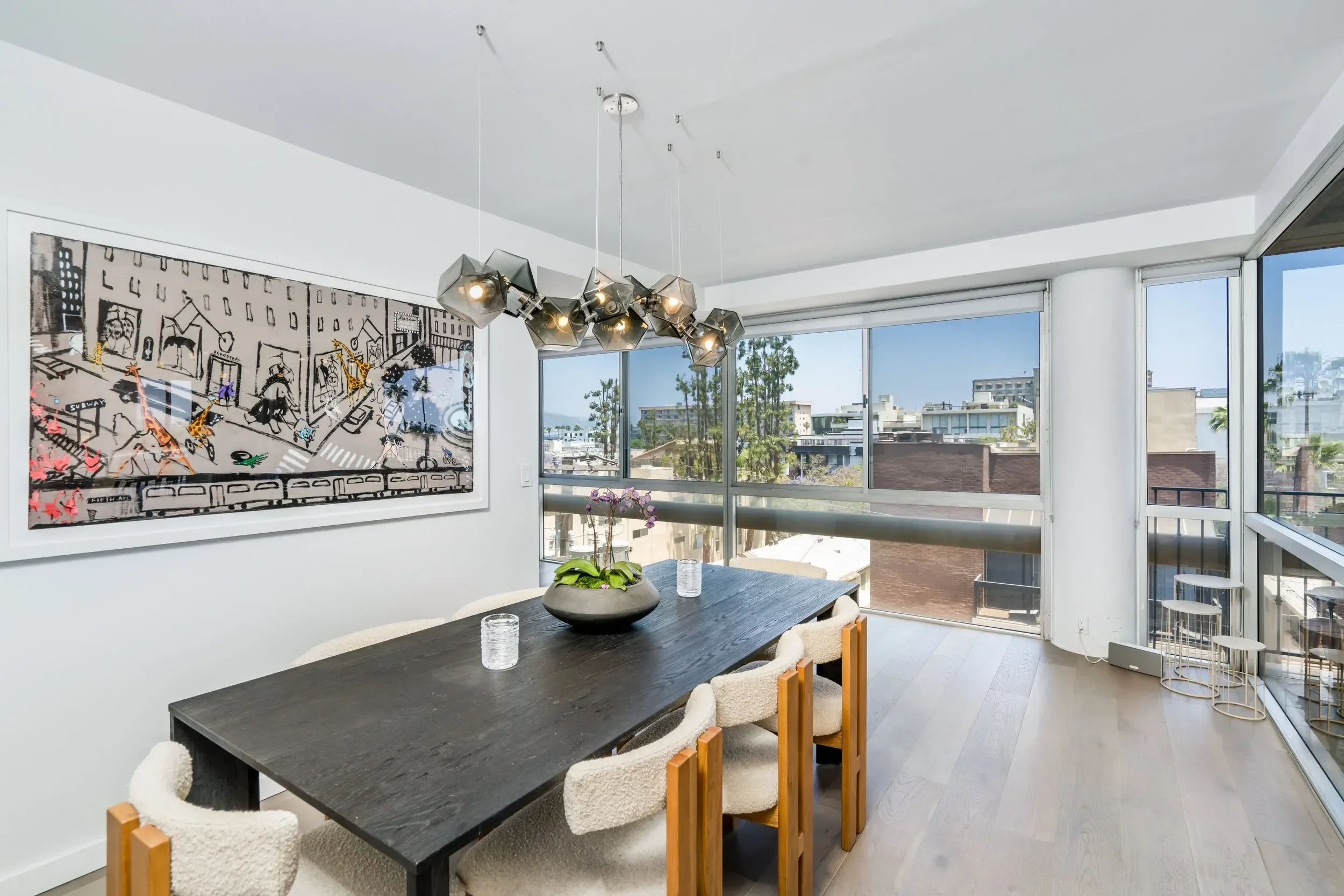 Modern dining area with black wooden table, six cushioned chairs, white walls, large city view window, abstract wall art, chandelier, and potted plant on table.