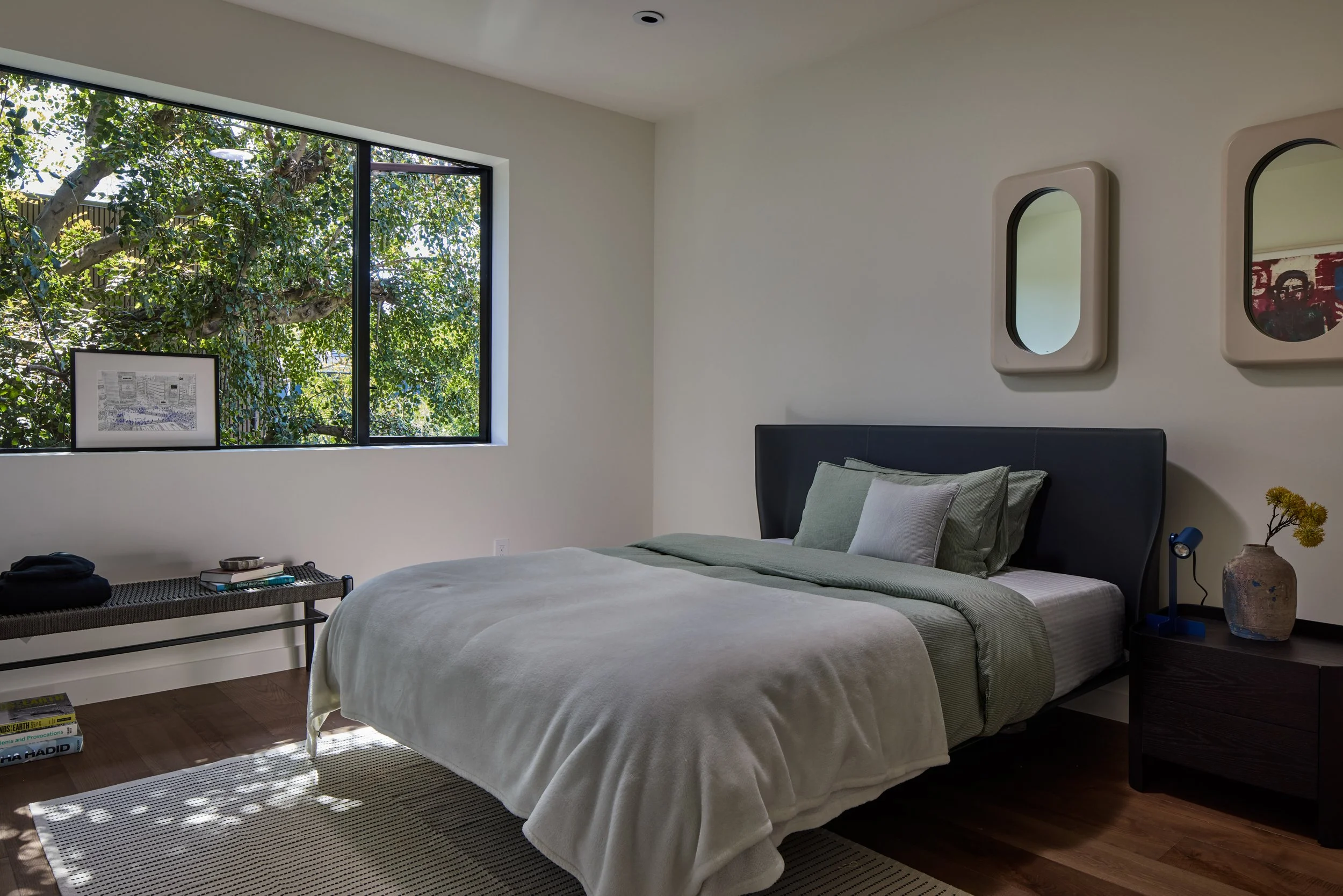 A modern bedroom with a large window letting in natural light and revealing green trees outside. The room has a bed with gray and white bedding, a black headboard, two oval mirrors on the wall, a side table with a decorative vase, a black desk, and s