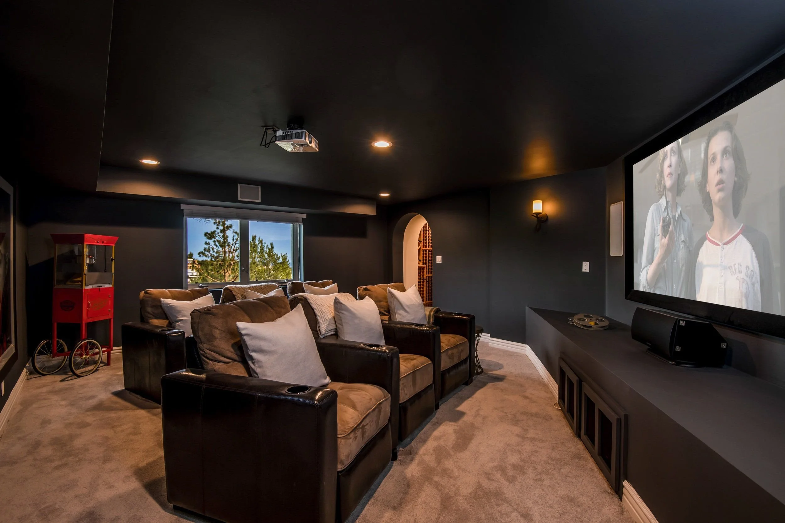A home theater room with a large screen showing two women, a dark ceiling with recessed lighting, a window with trees outside, beige sofas with pillows, a red popcorn machine, and a ceiling-mounted projector.