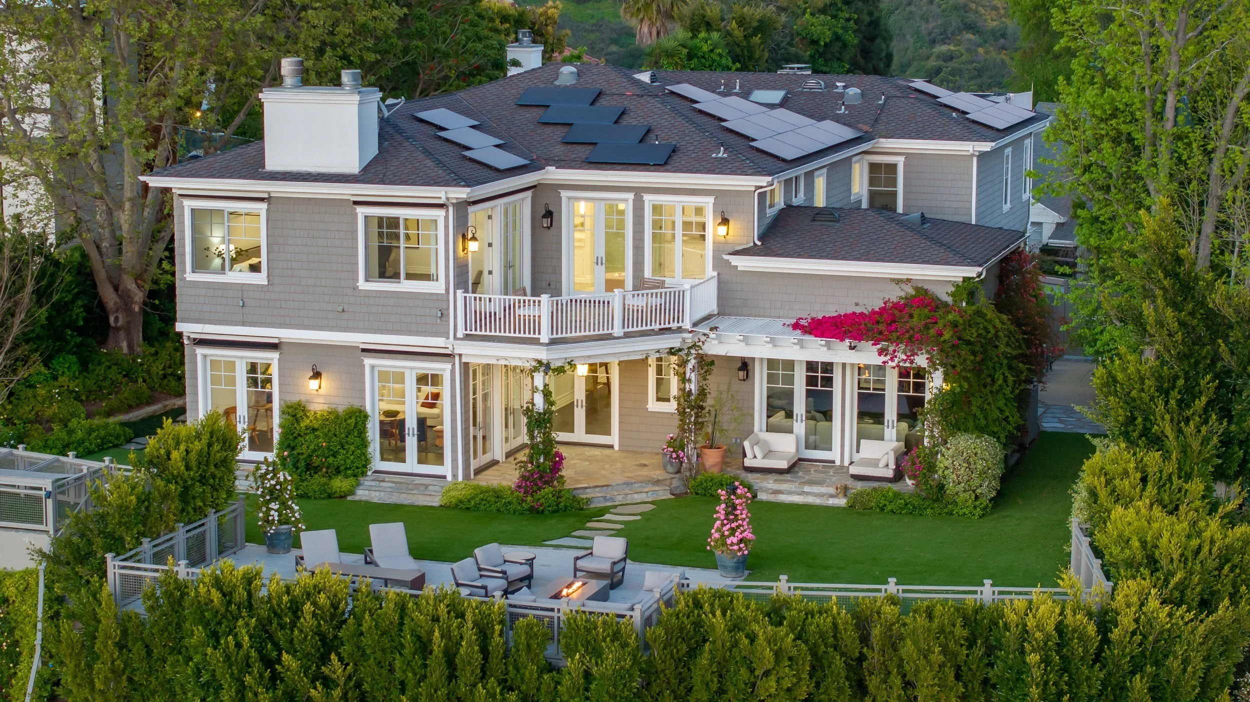 An aerial view of a two-story house with grey siding, multiple windows, and a deck with outdoor seating. The roof has solar panels installed on it. The backyard features a patio with a fire pit, a lawn, flowering plants, and surrounded by lush green 