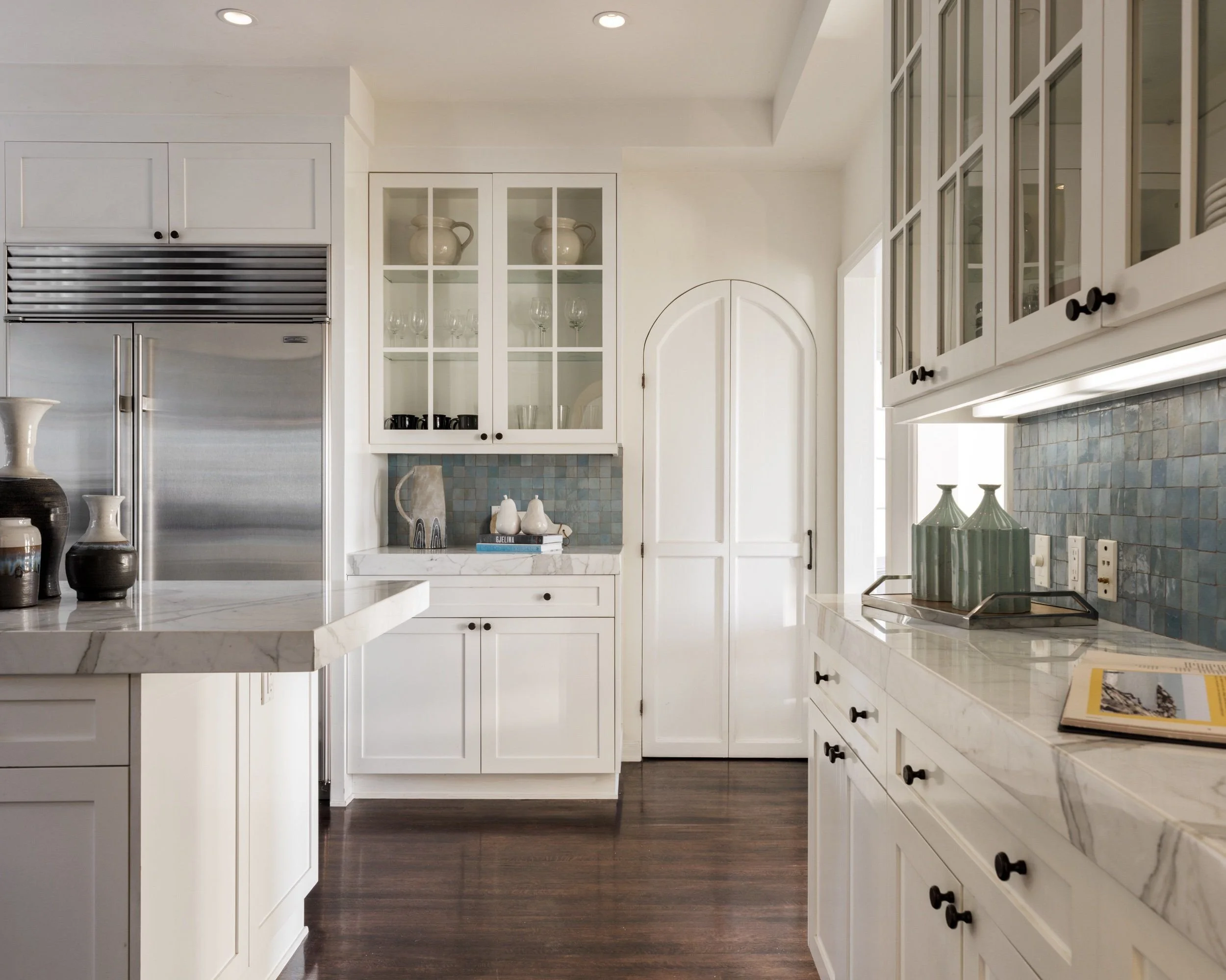 White kitchen with cabinets, a built-in refrigerator, decorative vases, and glassware, with a marble countertop and dark hardwood floors.