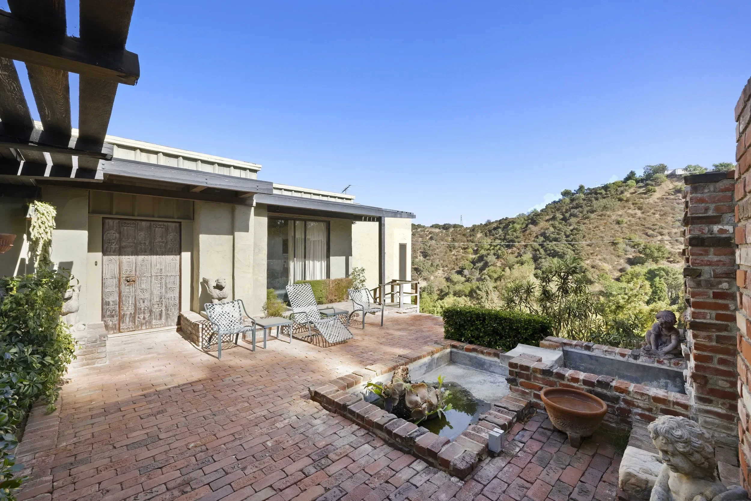 Backyard patio with brick flooring, outdoor chairs, plants, and a small pond, overlooking hills under clear blue sky.