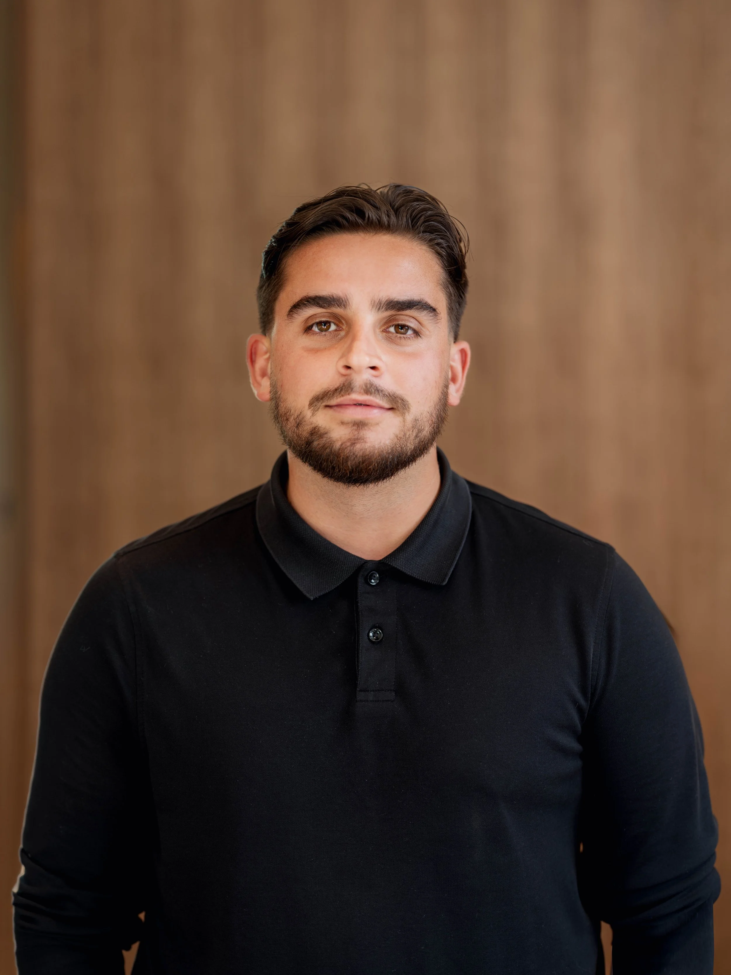 A young man with brown hair and a beard wearing a black collared shirt, standing against a blurred tan background.