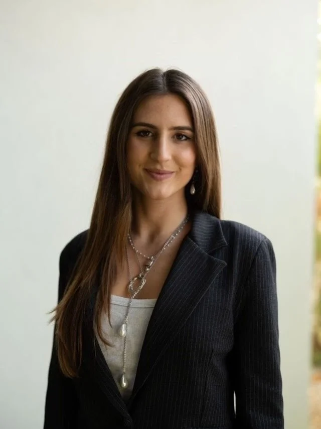 A woman with long, auburn hair and brown eyes, wearing a black sheer blouse with lace details, standing against a wooden background.