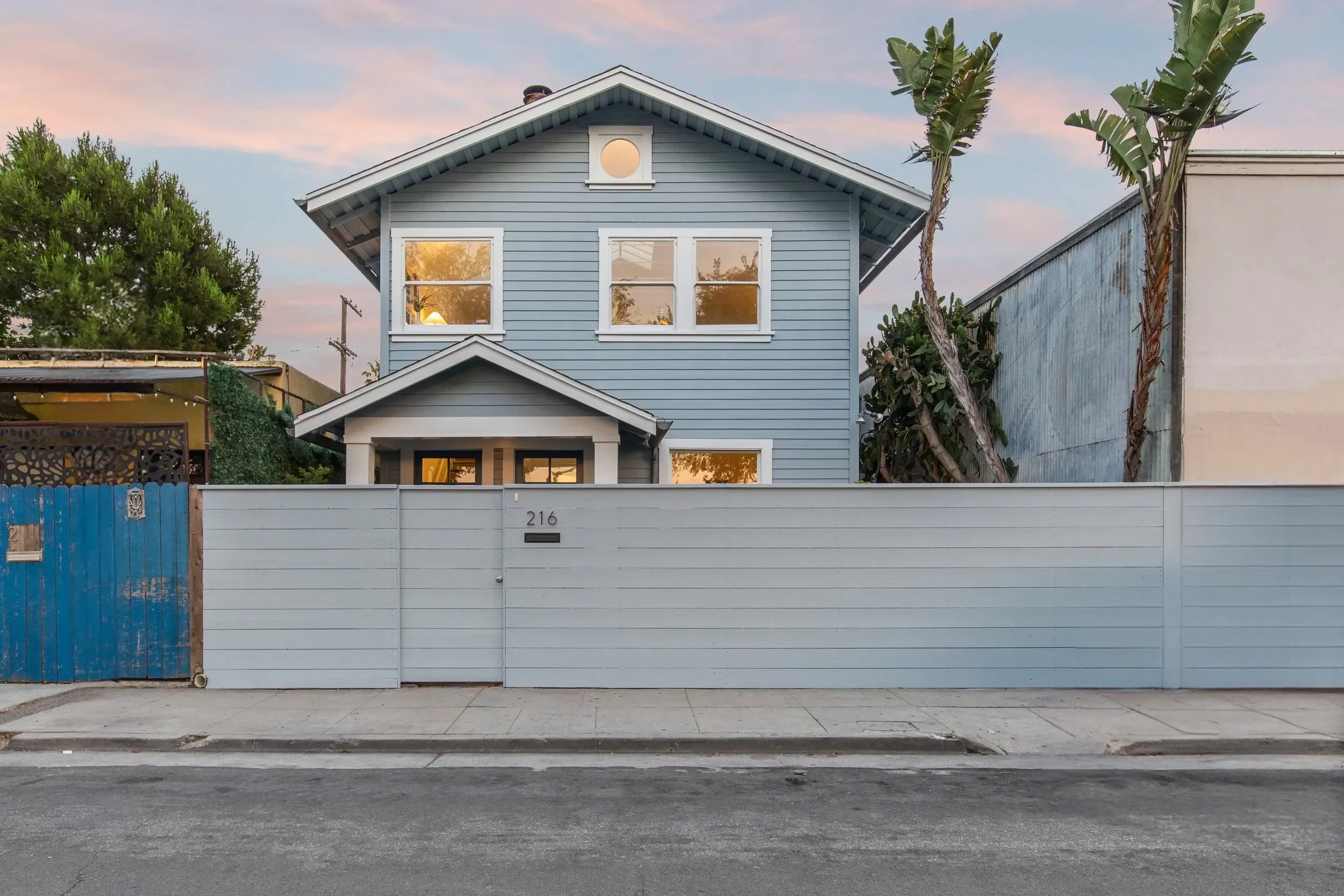 A two-story house painted light blue with white trim, behind a high white fence with house number 216, situated on a street with a sidewalk and gray pavement, with a small blue gate on the left and trees on both sides, and a sky with pinkish clouds i
