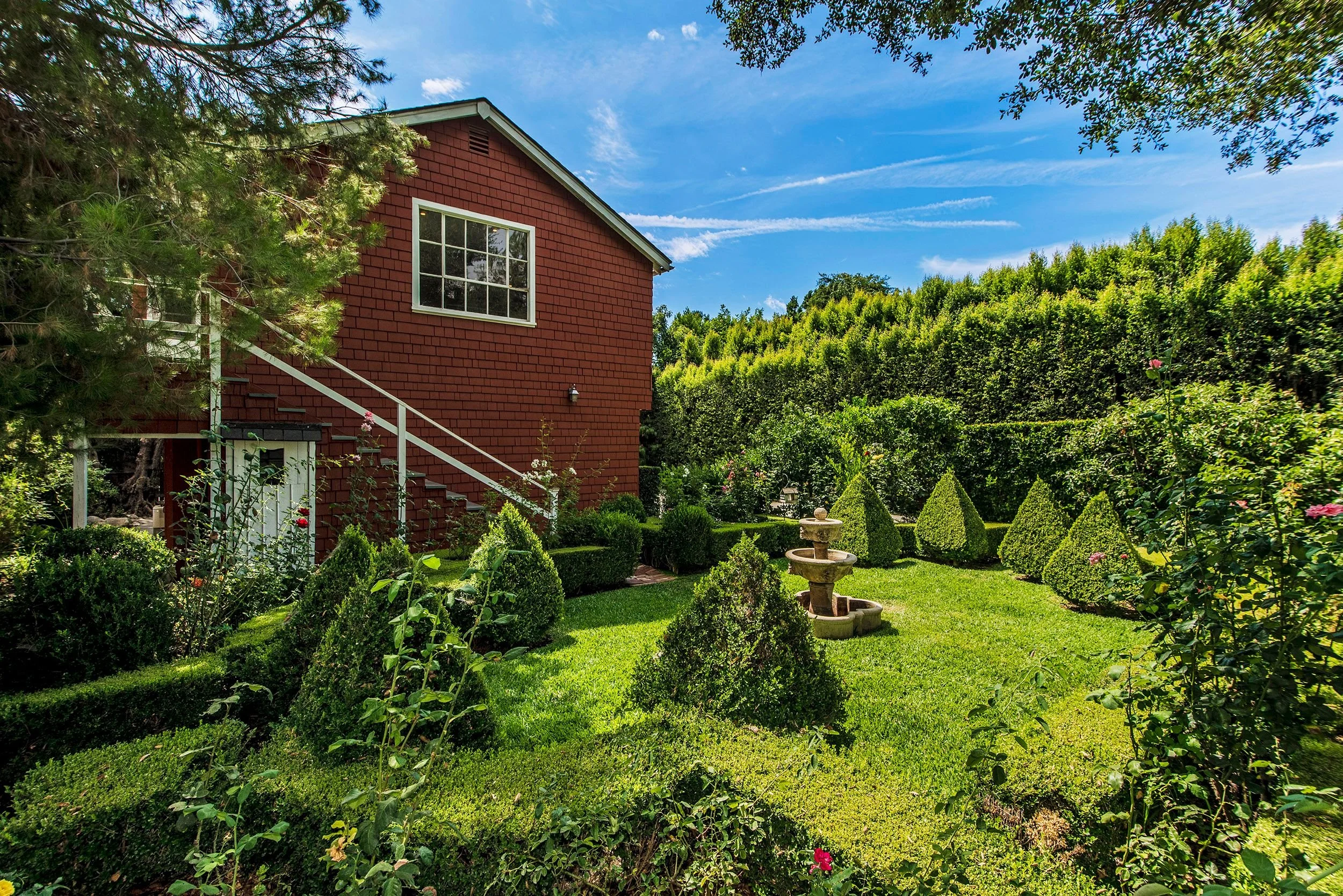 A red house with white trim and a large window on the upper level, surrounded by a lush green garden with bushes, trees, and a stone birdbath, under a partly cloudy sky.