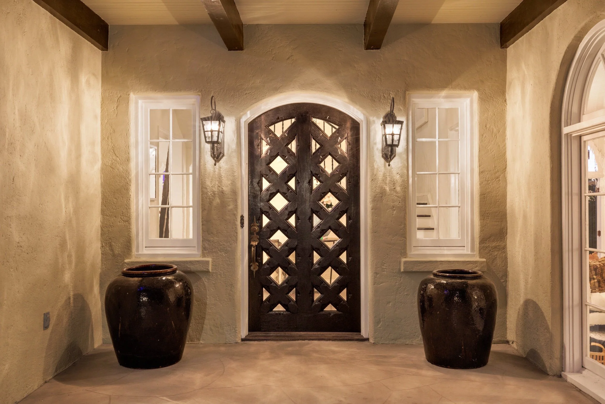 Entrance porch with a decorative wooden front door flanked by two windows, two large black ceramic vases on the floor, and wall lanterns on either side of the door, illuminated with warm lighting.