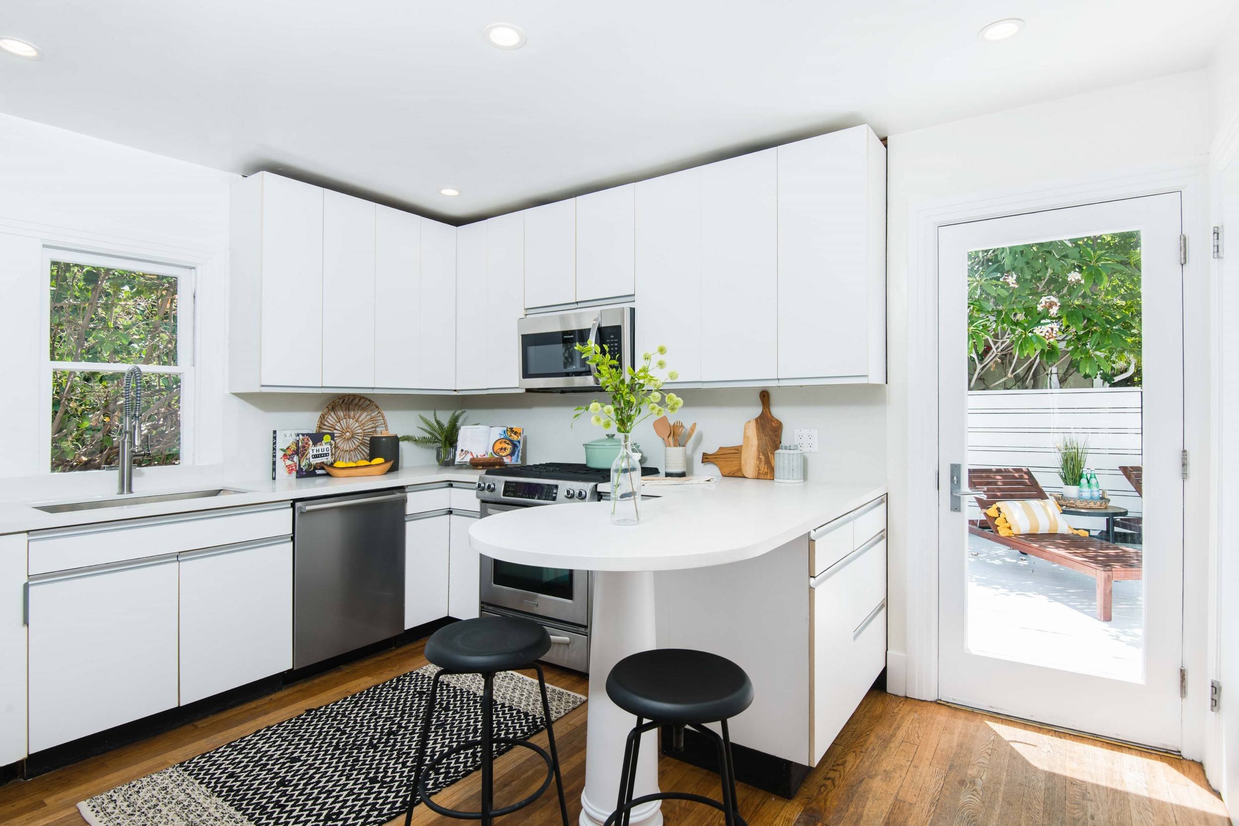 A modern kitchen with white cabinets, a black and white patterned rug, a small round white table with black stools, and an outdoor patio with chairs and a yellow pillow.