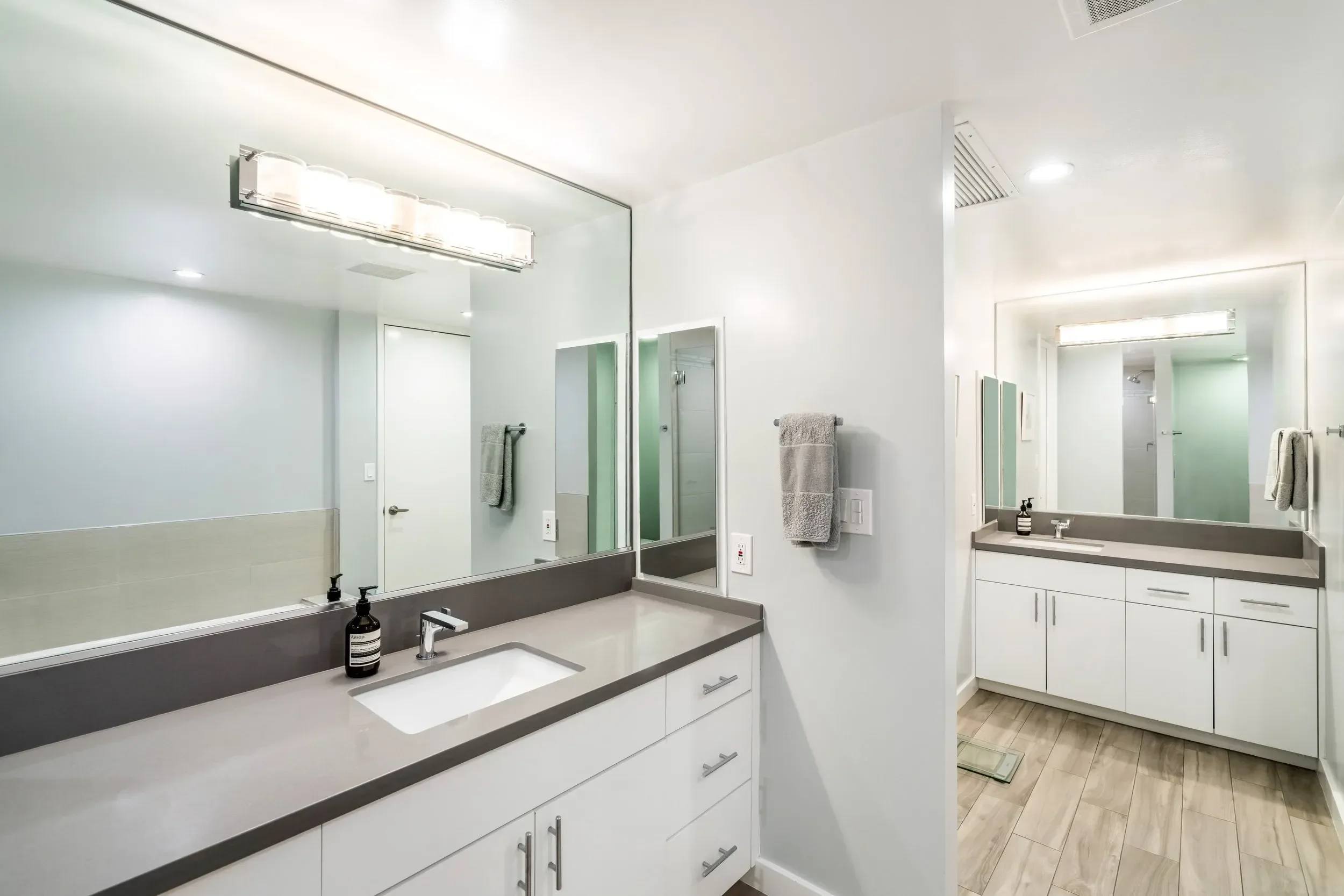A modern bathroom with a large mirror above a white countertop, two sinks, and white cabinetry. The bathroom has light-colored wood flooring, and there are towels hanging on the wall. There is a soap dispenser on the counter and a small bottle. The r