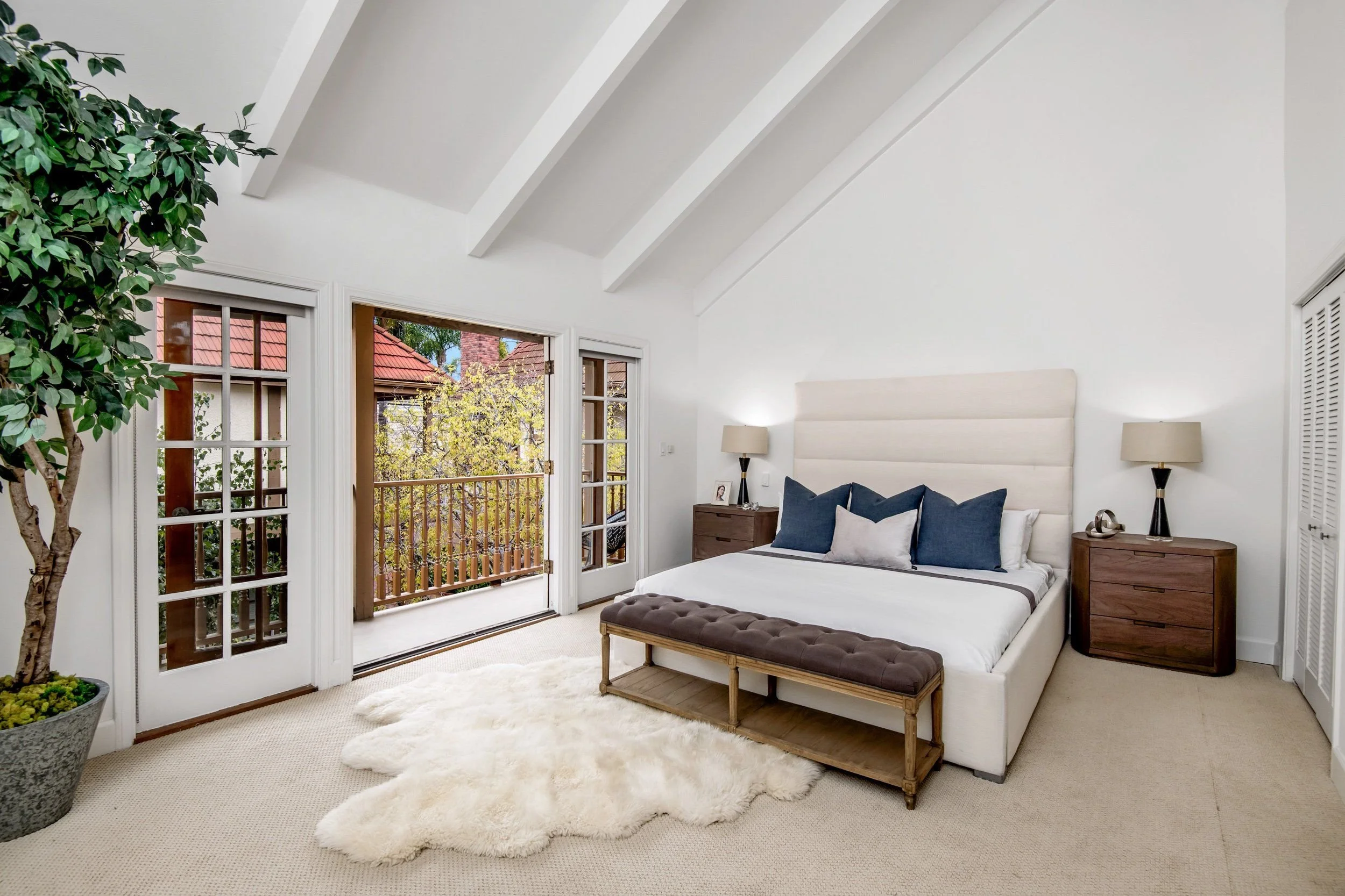 A bedroom with white walls and ceiling, two bedside tables with lamps, a bed with pillows, a wooden bench at the foot, a large green potted plant, and open French doors leading to a balcony with trees outside.