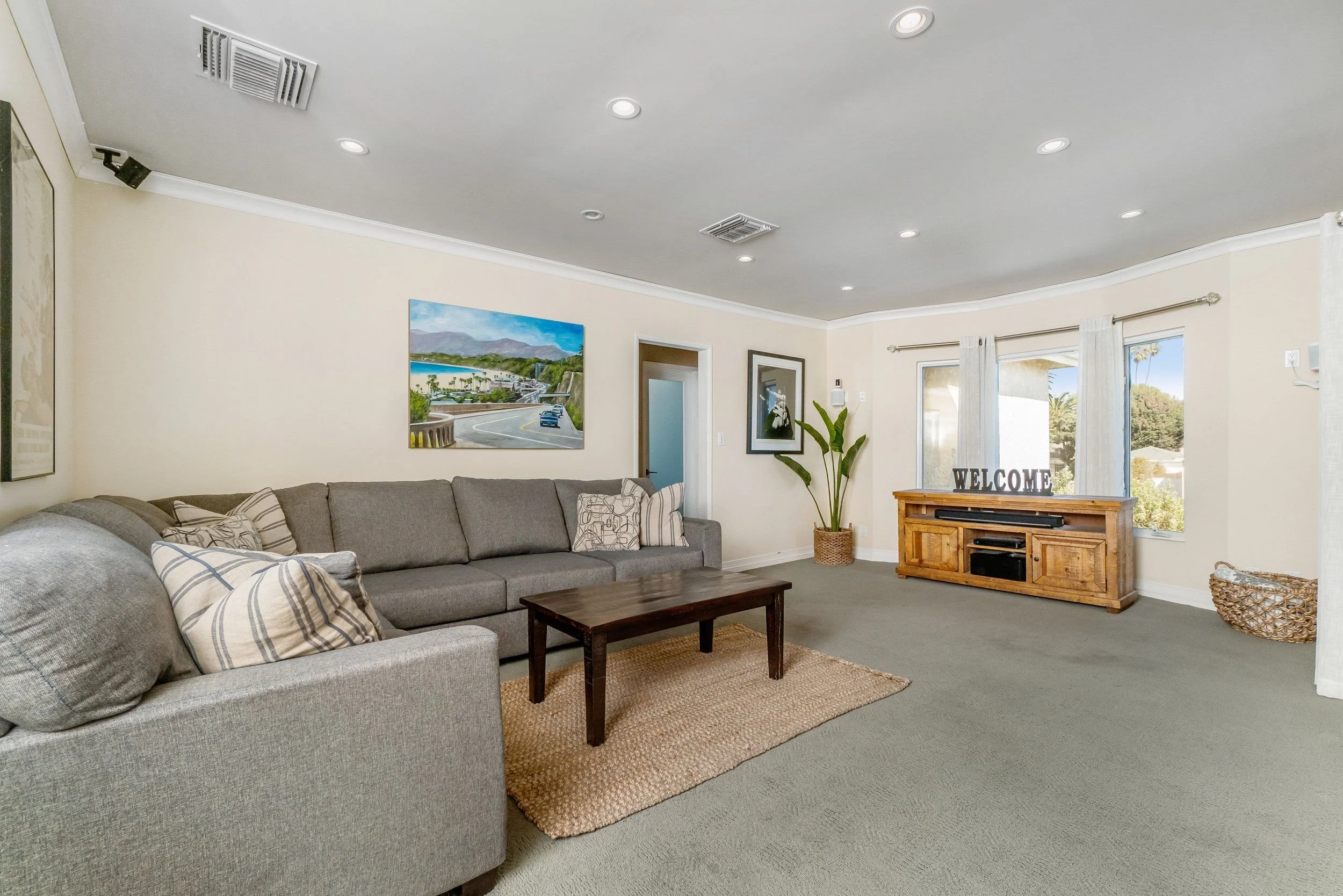 Living room with a gray sofa, wooden coffee table, potted plant, wall art, and a wooden media console with a 'WELCOME' sign. Large windows with white curtains.