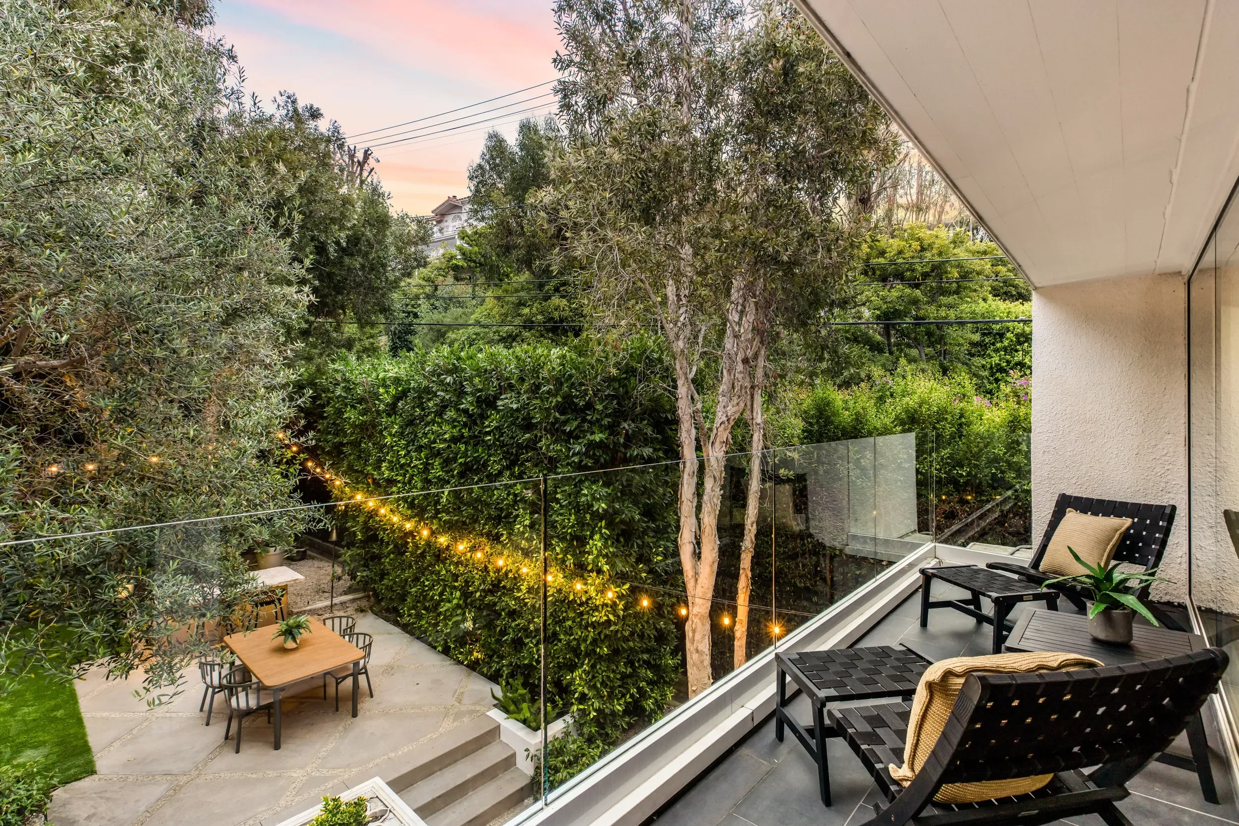 A backyard patio area with outdoor seating, lush greenery including trees and hedges, string lights, and a neighboring house visible in the background, viewed from a balcony with glass railing.