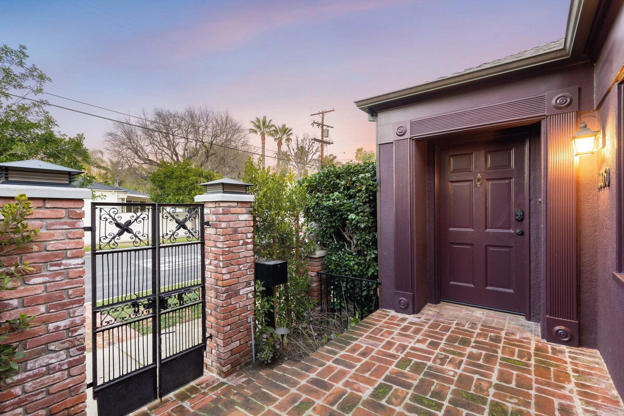 A brick walkway leading to a front door with an outdoor wall light, a black wrought iron gate, brick columns, green bushes, trees, utility poles, and power lines under a sunset sky.