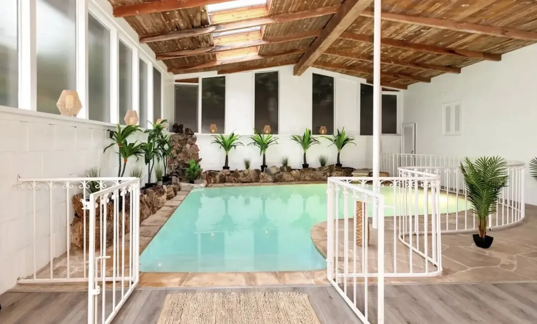 Indoor swimming pool with white safety gate, surrounded by potted plants, rocks, and a wooden ceiling with skylights.