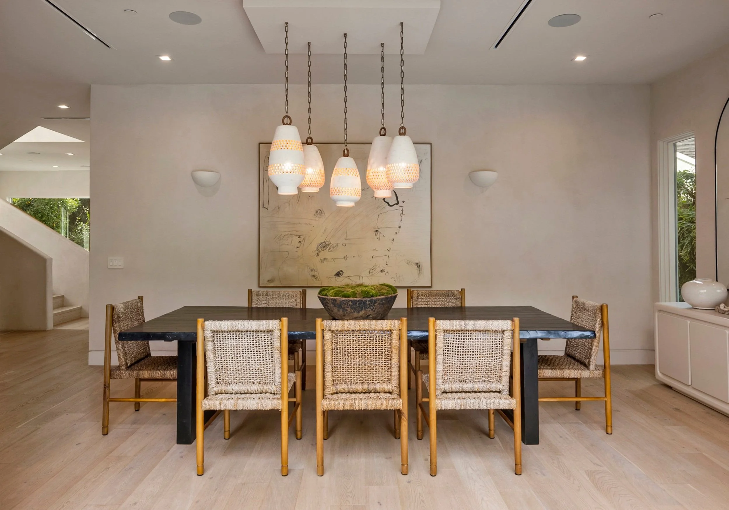 Modern dining room with a black table, woven dining chairs, and a bowl of moss centerpiece. Five pendant lights hang above the table, and a large abstract painting is on the wall behind. Windows and a staircase are visible in the background.