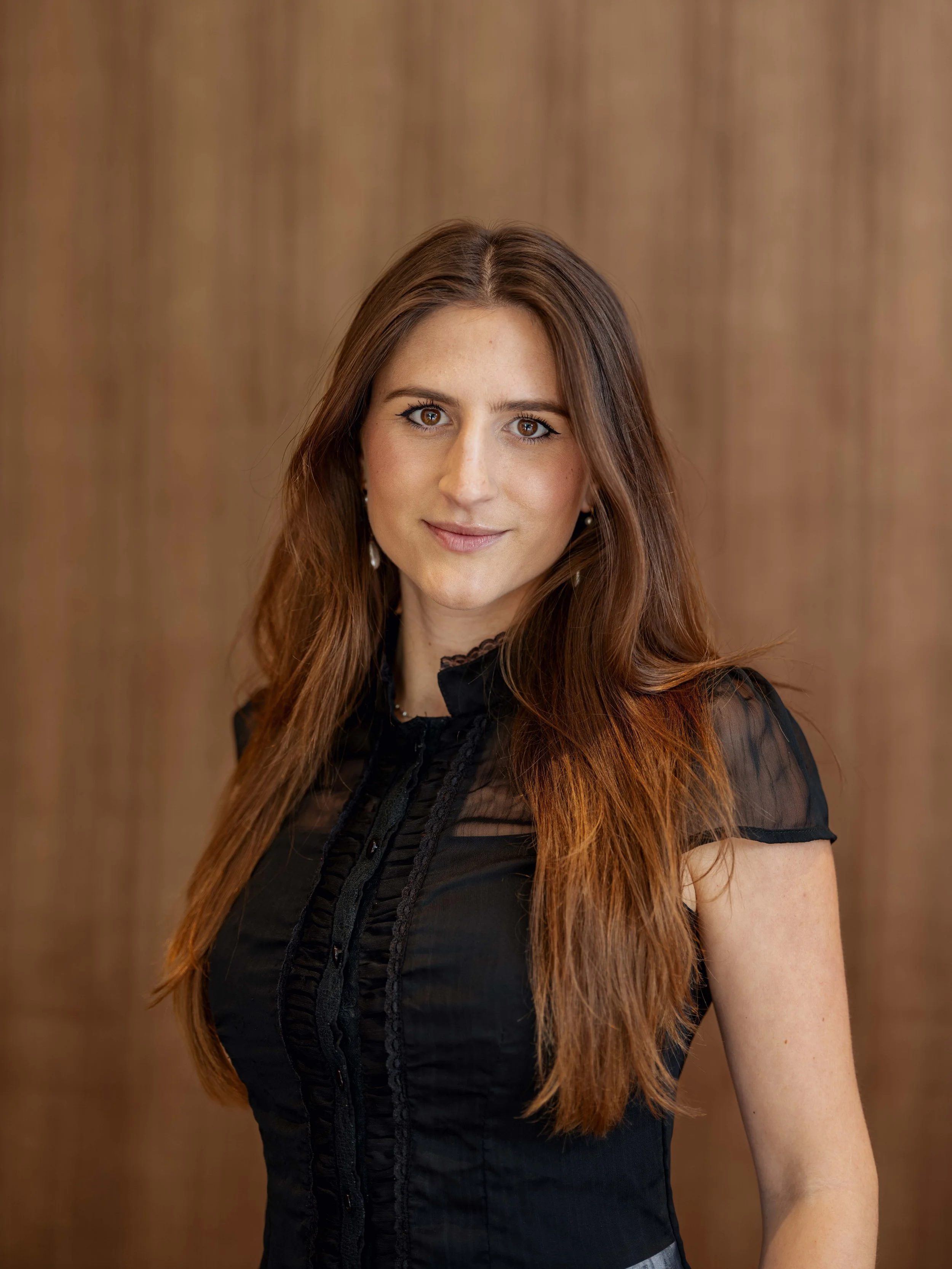A woman with long, auburn hair and brown eyes, wearing a black sheer blouse with lace details, standing against a wooden background.