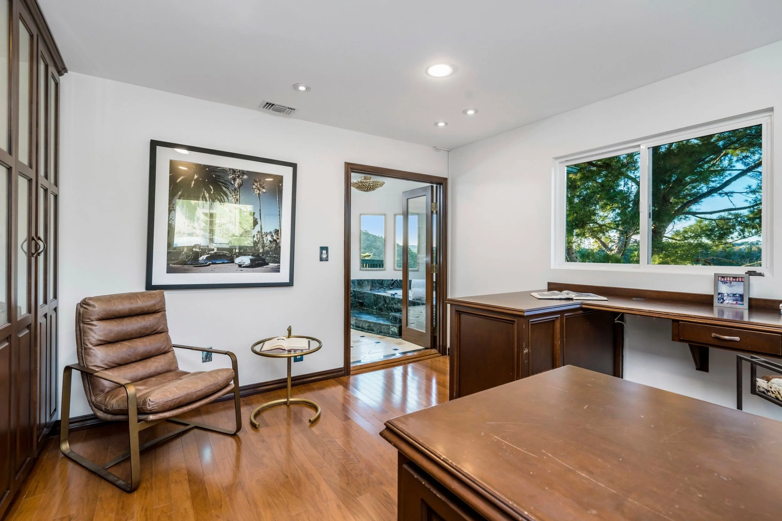 Interior of an office with a wooden desk, a leather chair, framed photos and artwork on white walls, a large window showing green trees outside, and a door leading to a bathroom.