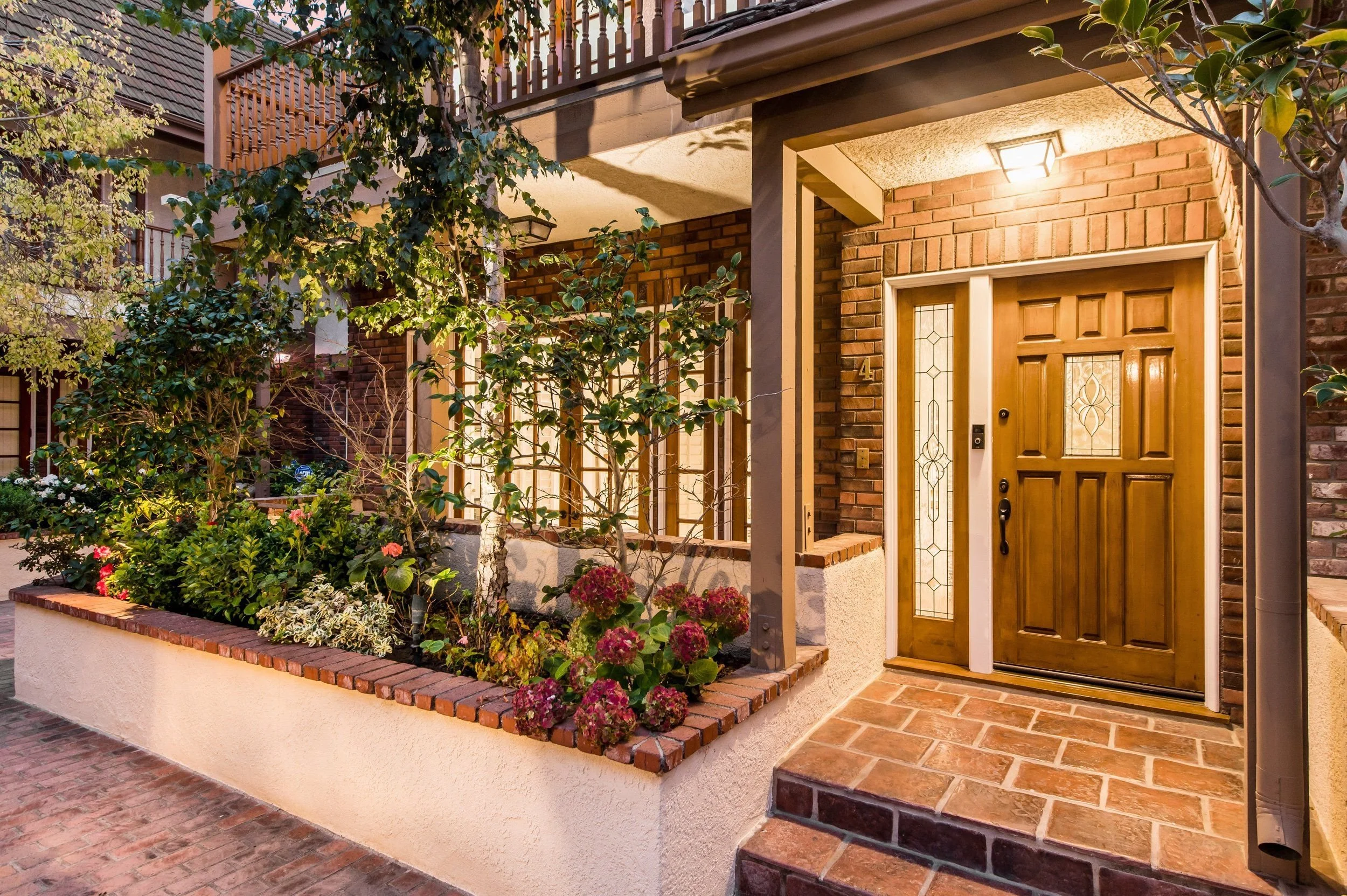 Brick house entrance with wooden door and stained glass window, surrounded by trees, plants, and flowers, with a porch light on in the evening.