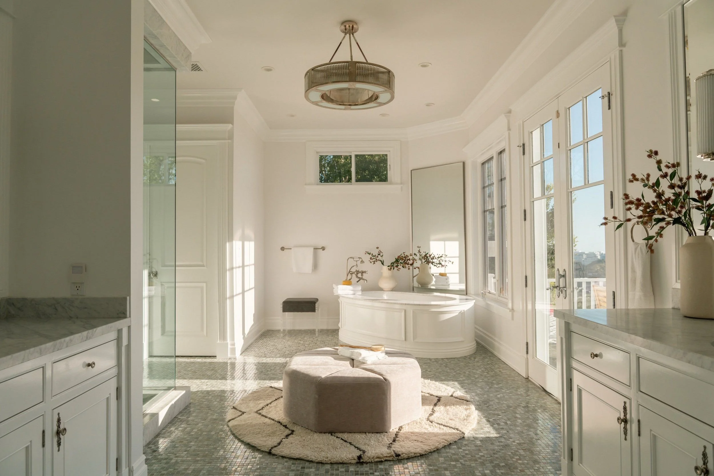 Bright bathroom with large windows, white cabinetry, a round beige ottoman, and decorative vases and flowers.